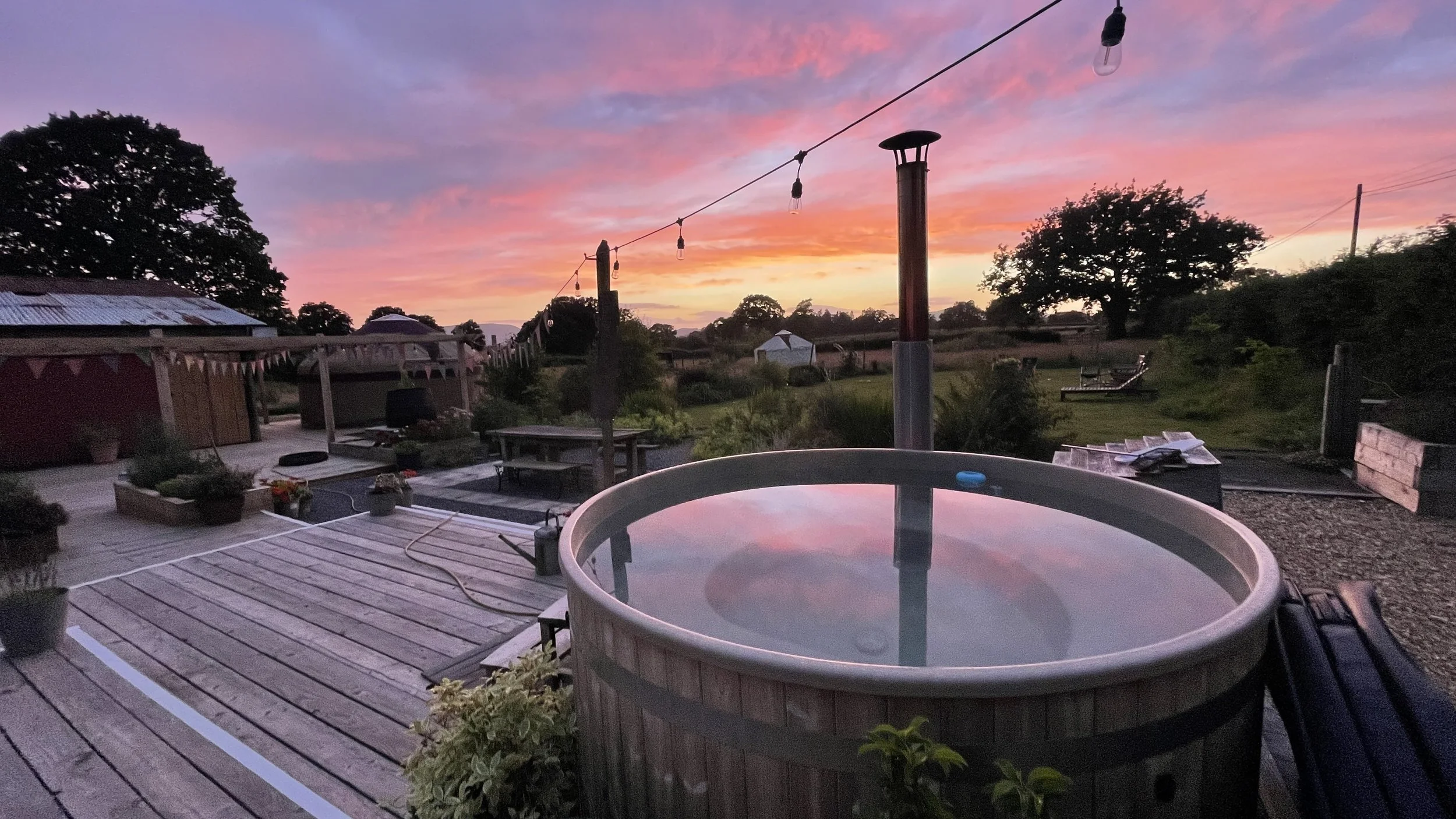 An outdoor deck with a hot tub reflecting a colorful sunset sky, surrounded by trees, plants, and outdoor furniture.
