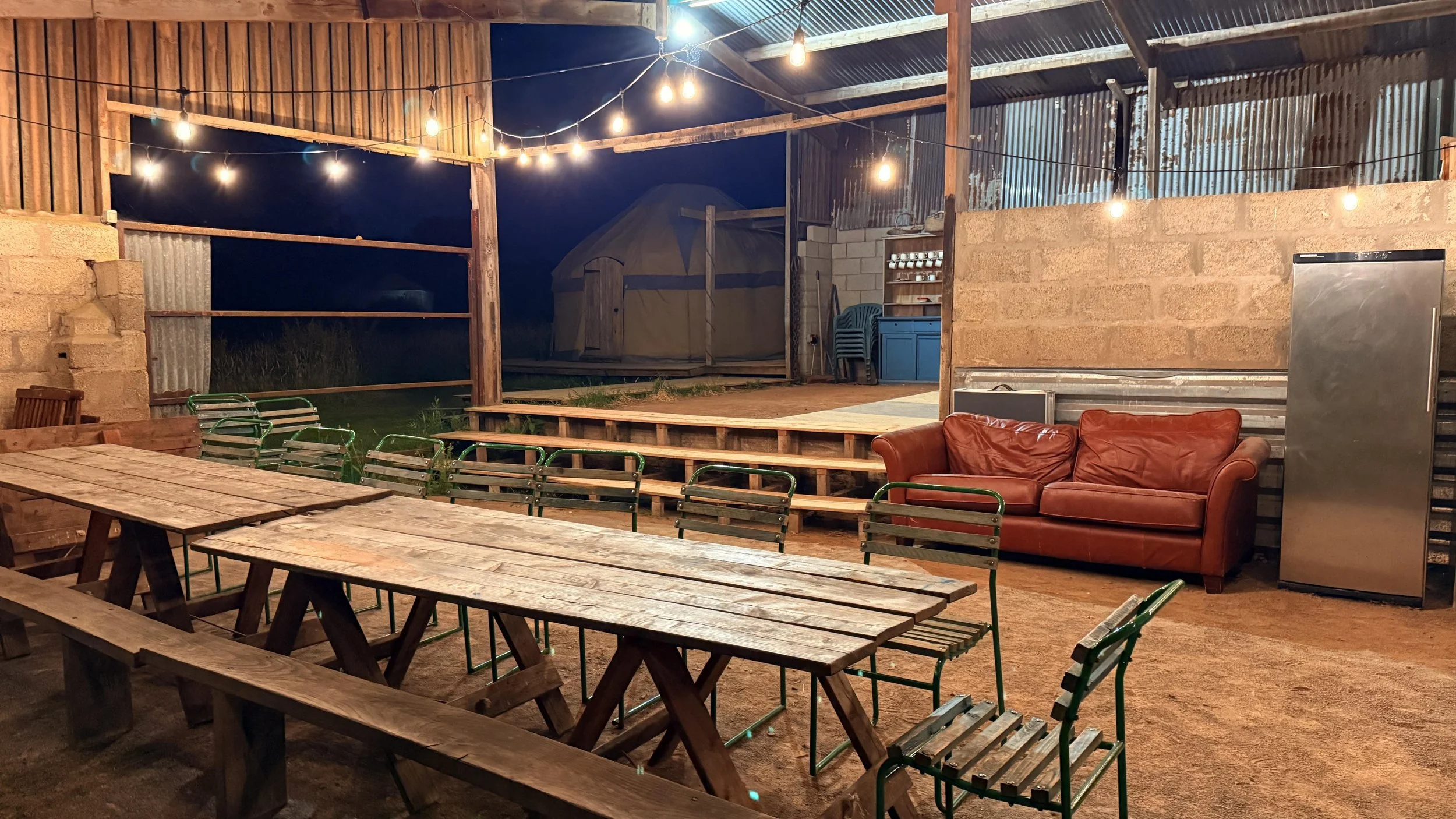 Indoor rustic dining area with string lights, wooden tables, green metal chairs, a brown leather couch, a stainless steel refrigerator, and a raised outdoor stage with chairs and a tent in the background.