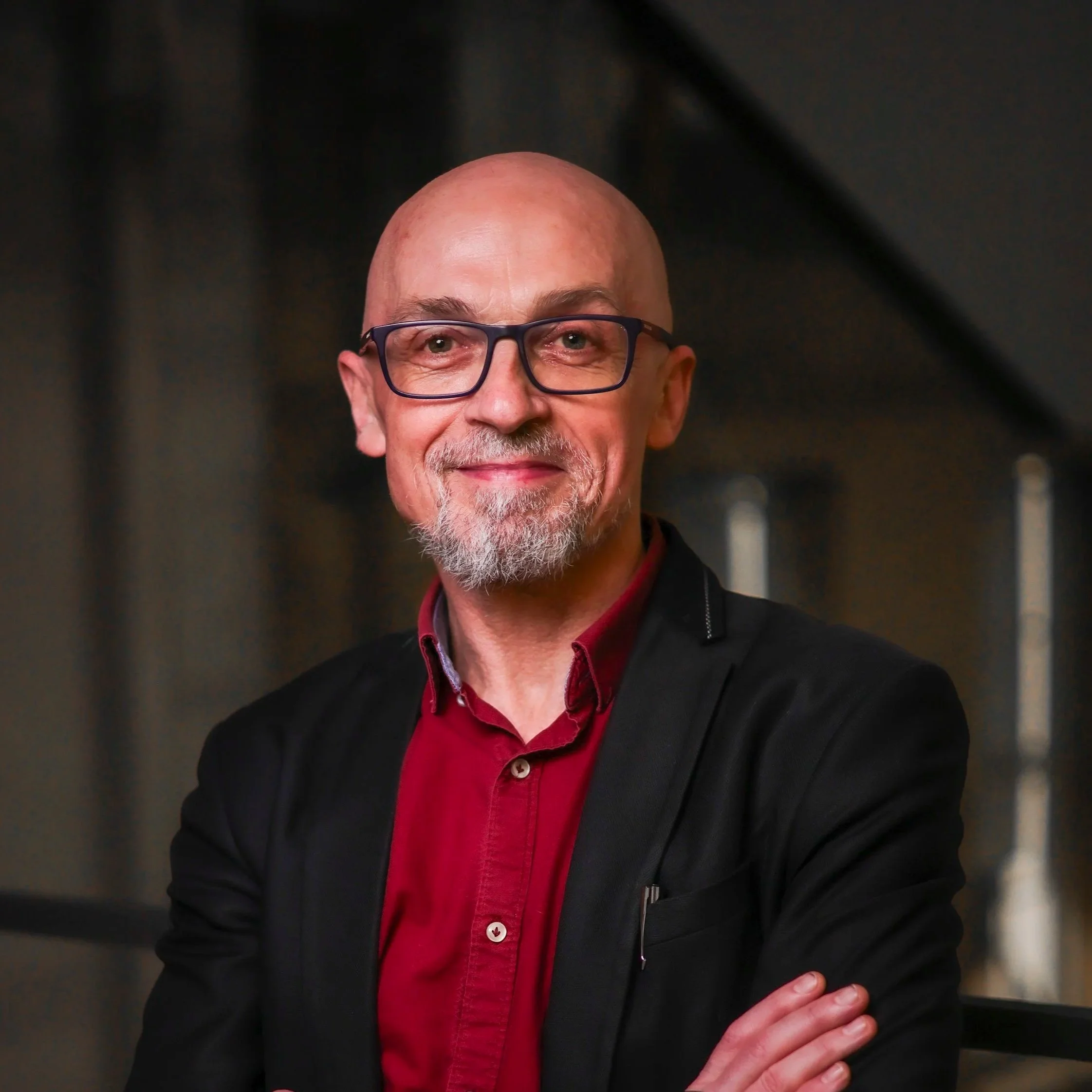 A middle-aged man with glasses and a beard, wearing a blue collared shirt, standing in front of a plain white wall.