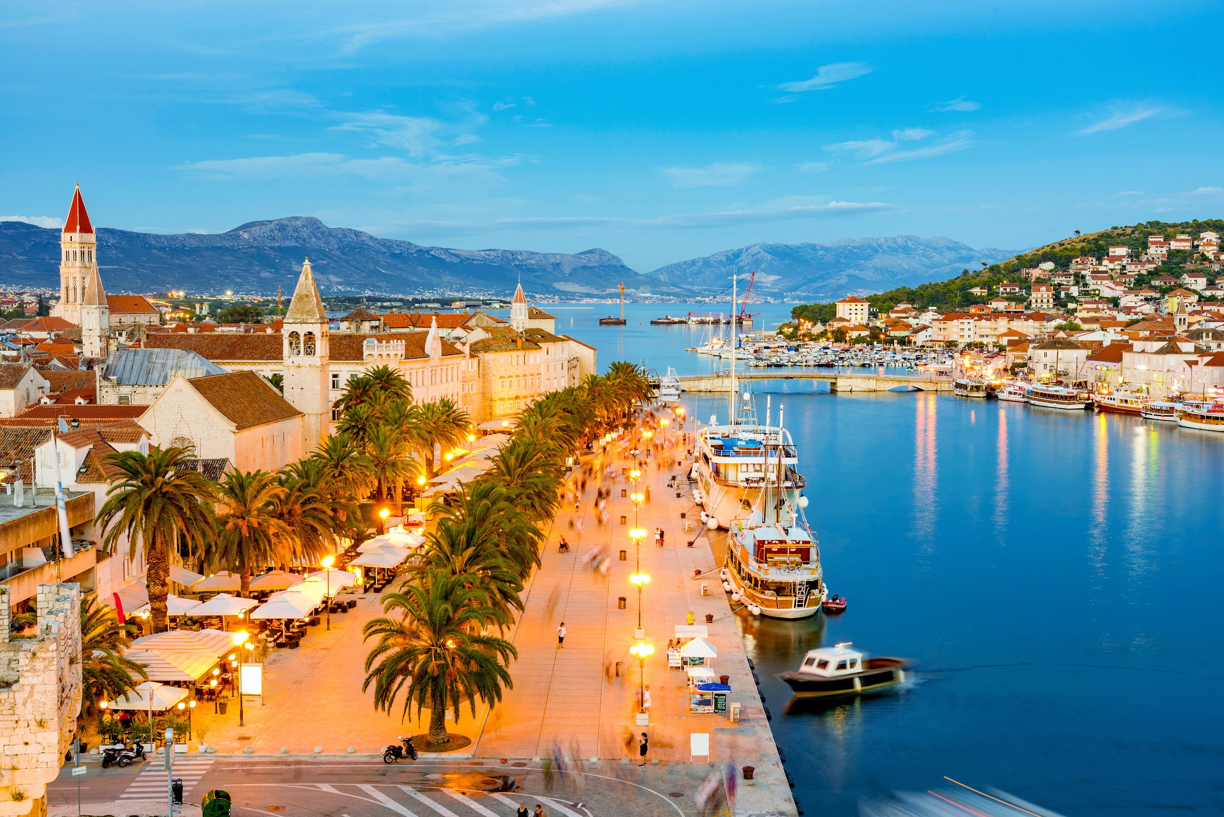 A coastal city promenade at dusk with palm trees, outdoor cafes, and boats docked at the harbor, overlooking a calm bay with hills and mountains in the background.