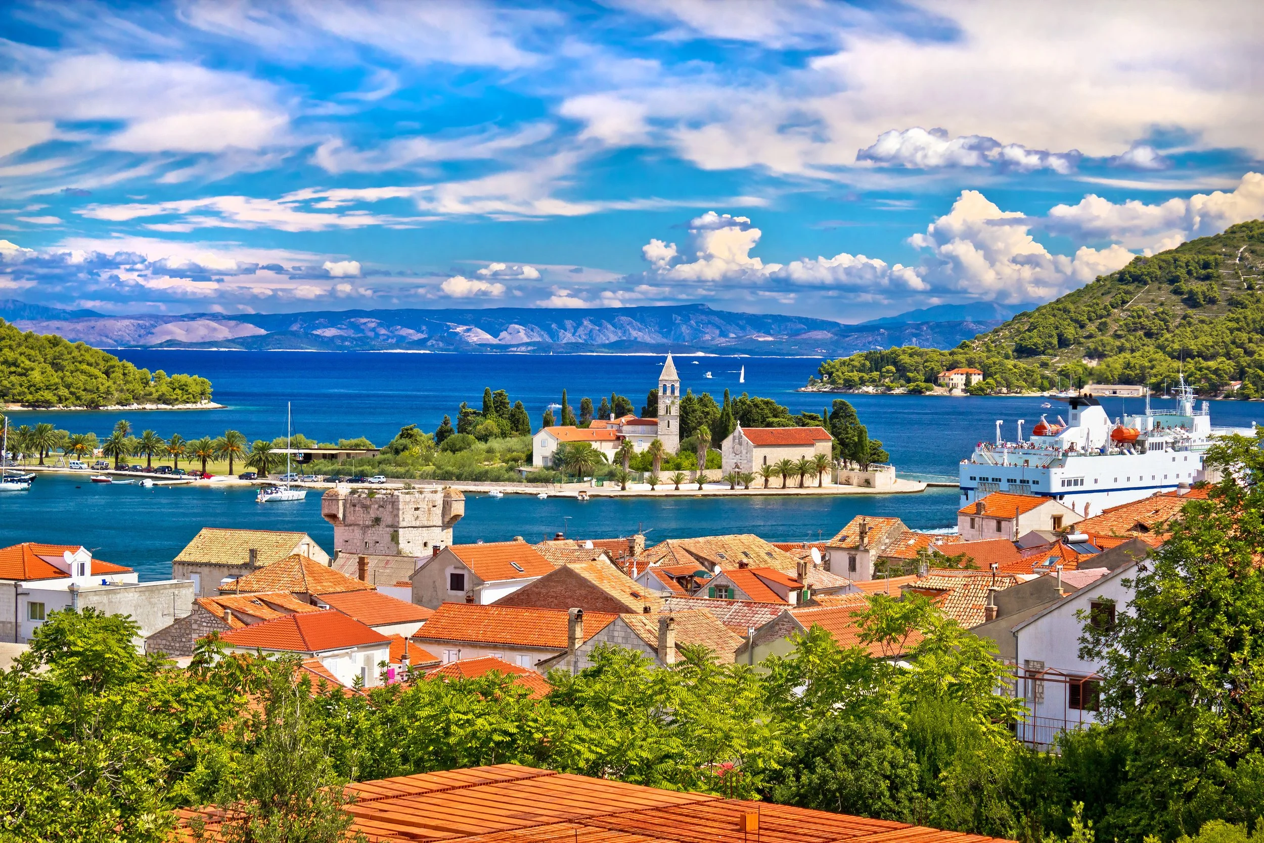 Scenic view of a coastal town with red-tiled roofs, a church with a tall steeple, a large white ship docked at the harbor, lush green trees, and mountains in the background under a partly cloudy sky.