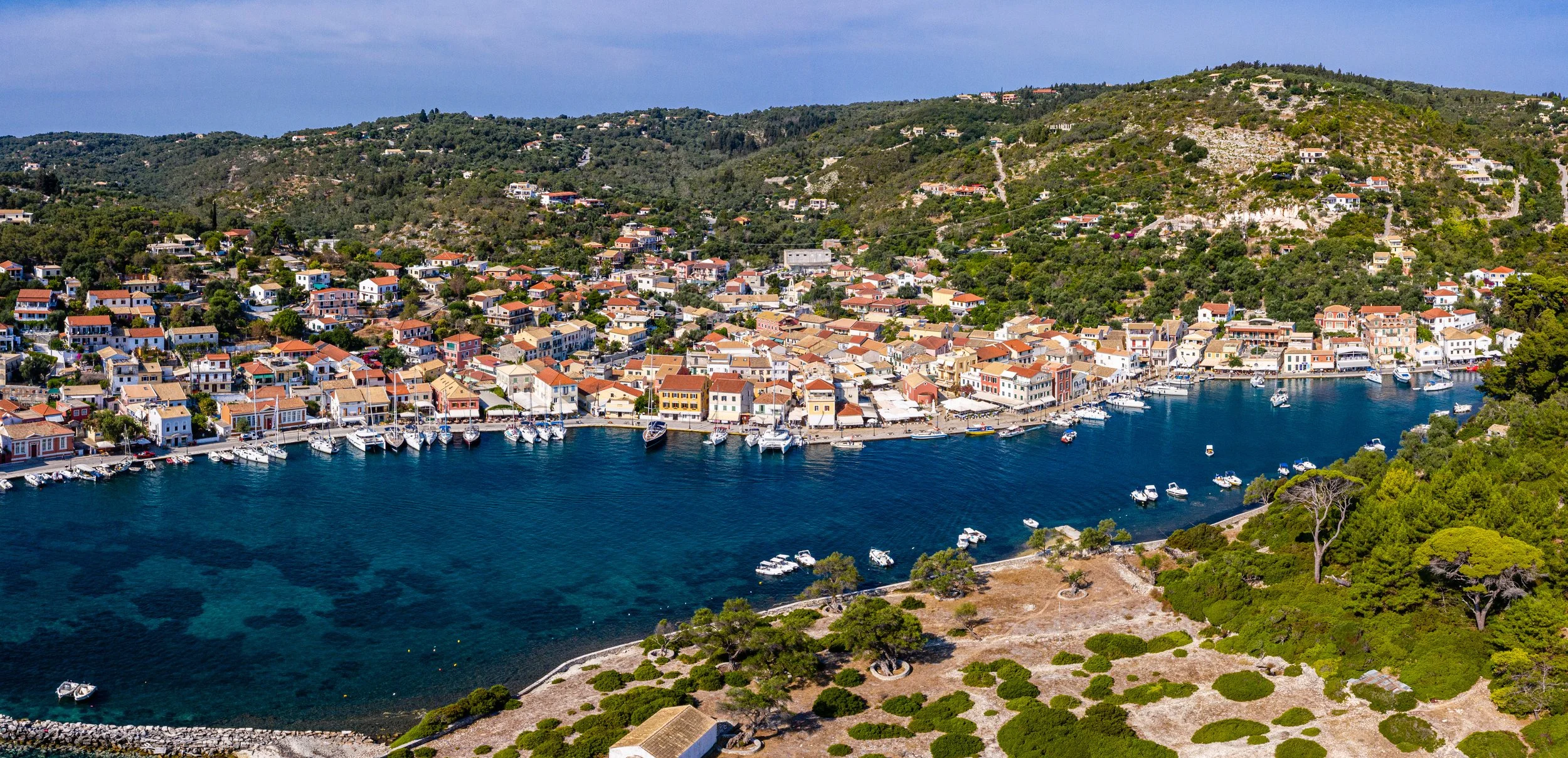 Aerial view of a coastal hillside town with colorful buildings, a marina with boats, and green hills in the background.