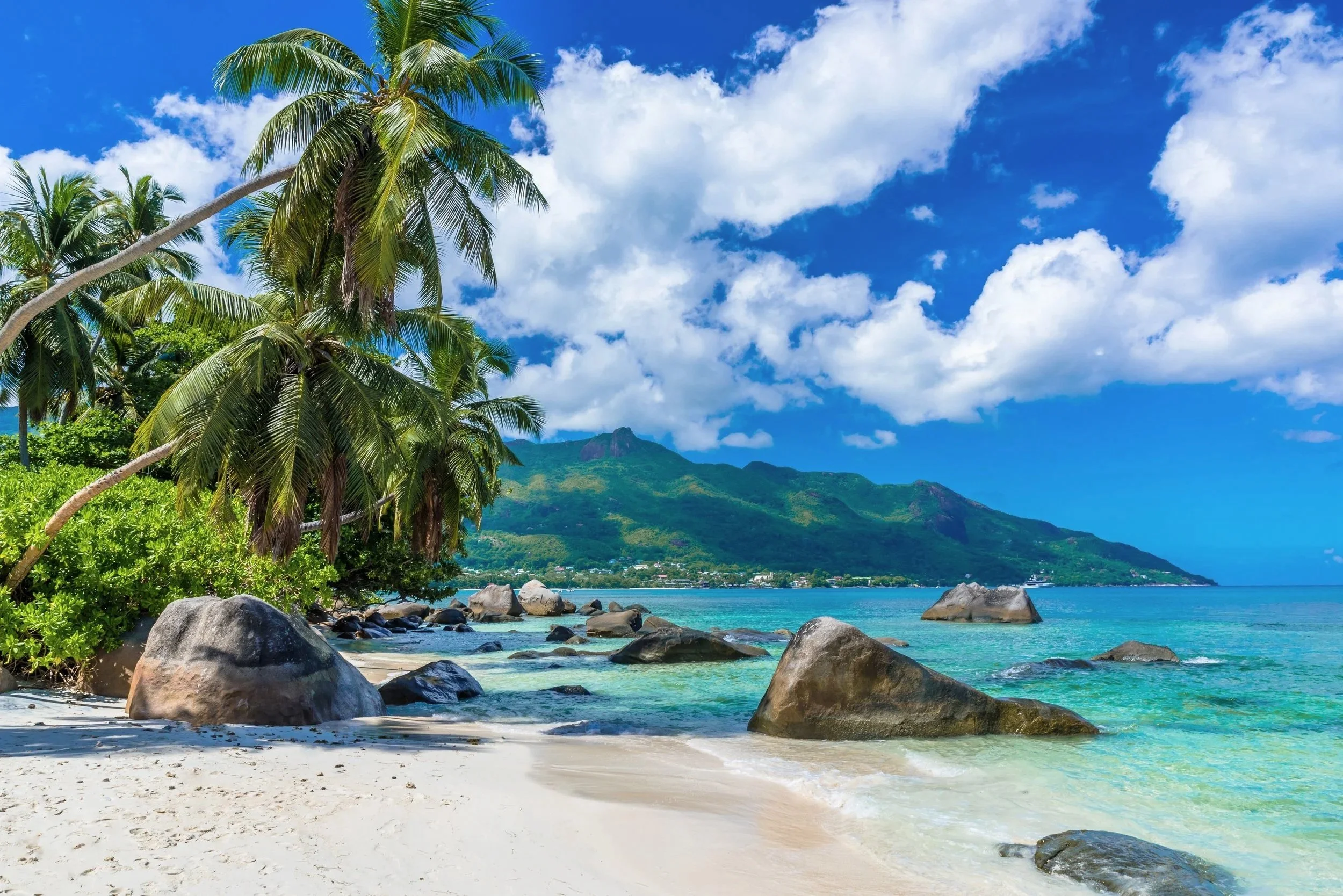 Tropical beach with white sand, large rocks, green palm trees, turquoise water, lush green mountains in the background, and a partly cloudy blue sky.