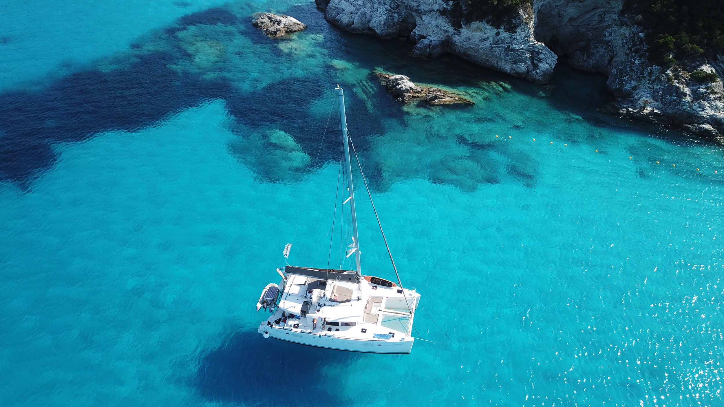 An aerial view of a white sailing boat floating in turquoise blue waters near rocky cliffs.