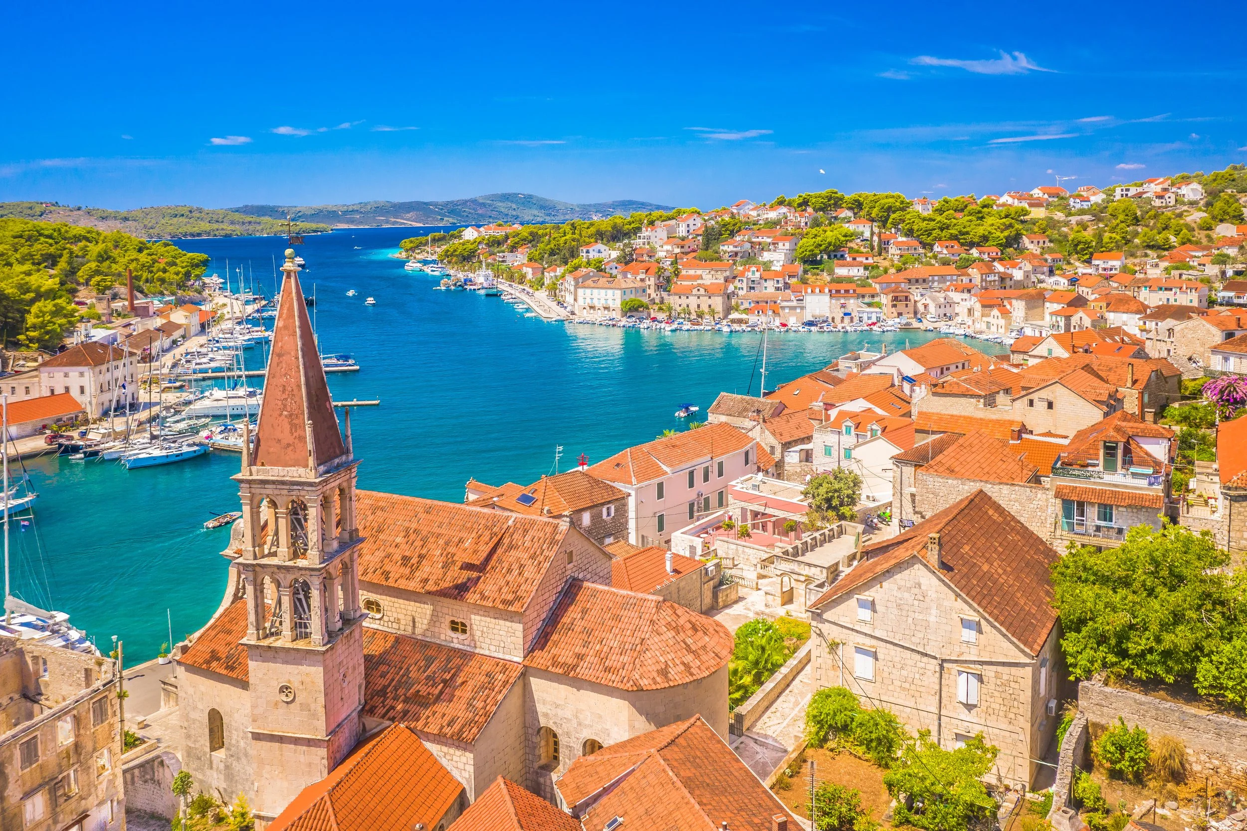 A coastal town with red rooftops, a church steeple, a marina with boats, and clear blue water under a partly cloudy sky.