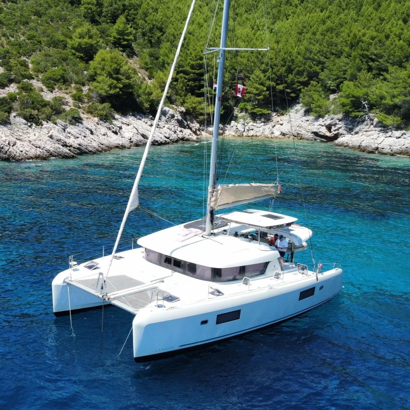 A white sailboat on clear blue water near a rocky shoreline with green trees, with flags flying from the mast.