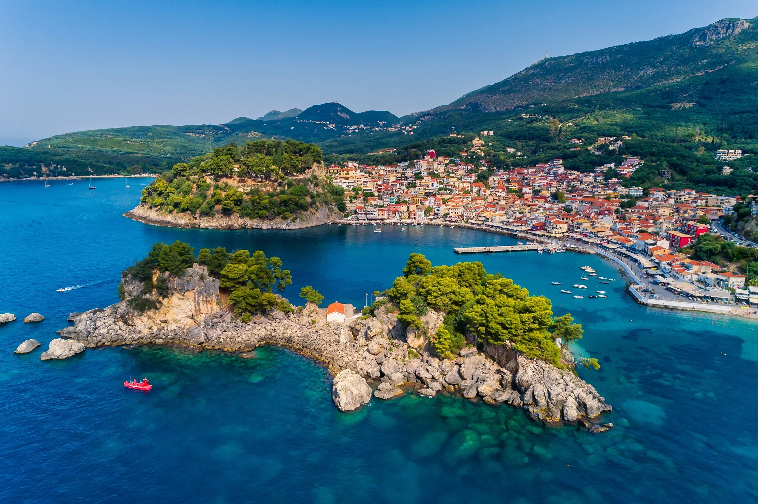Aerial view of a coastal town with colorful buildings, a harbor with boats, and green hills in the background on a sunny day.