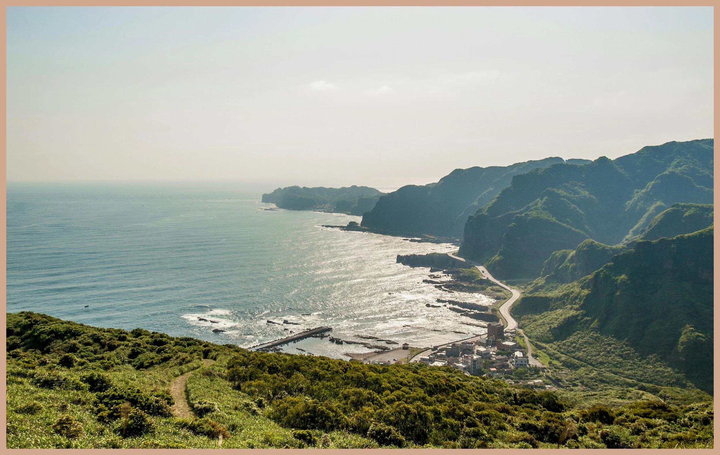 View of a coastal landscape with green mountains, a winding road, and the ocean under a clear sky.