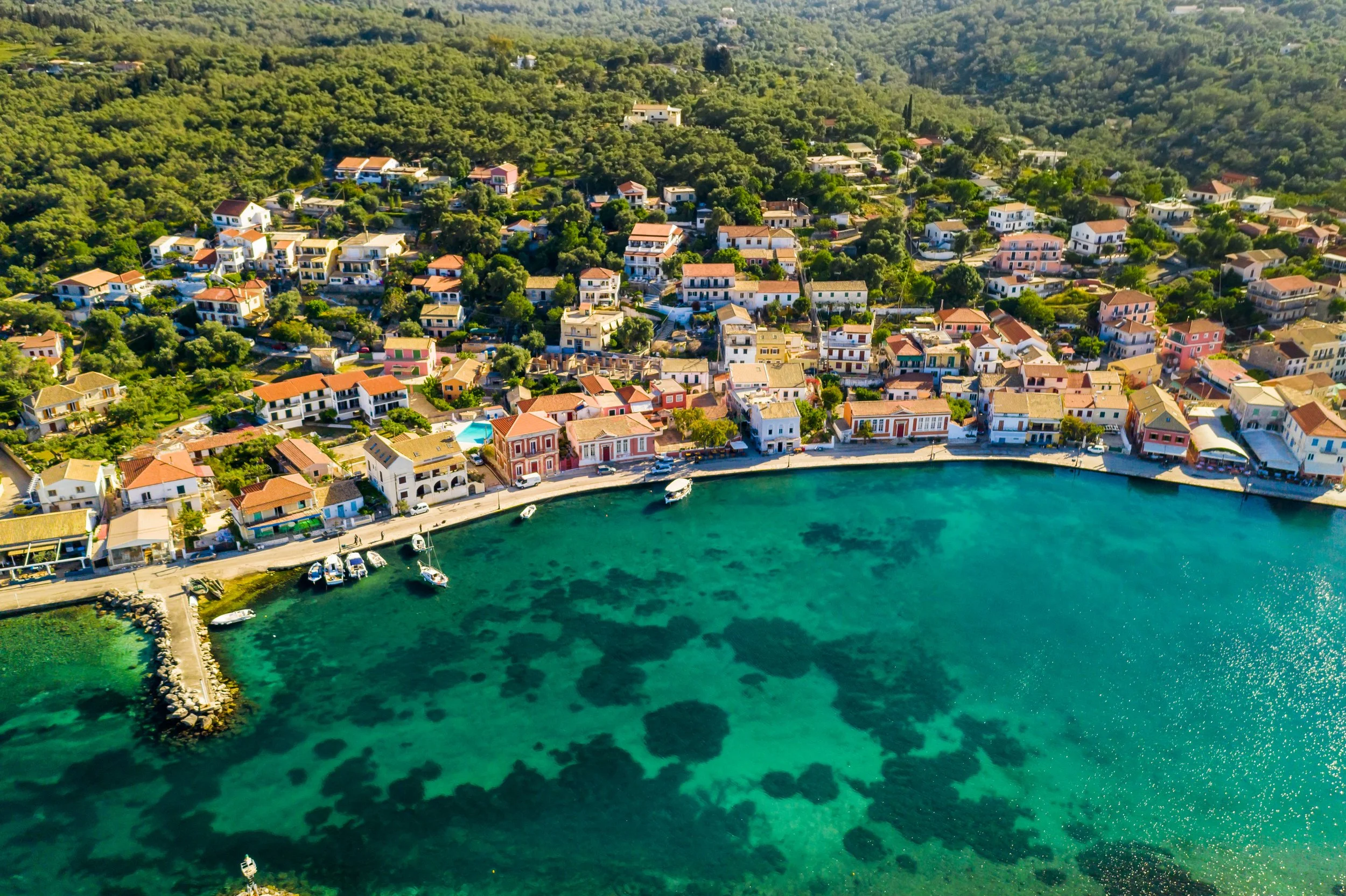 Aerial view of a Mediterranean coastal town with colorful buildings along a curved shoreline, clear turquoise water, and boats docked at the harbor.