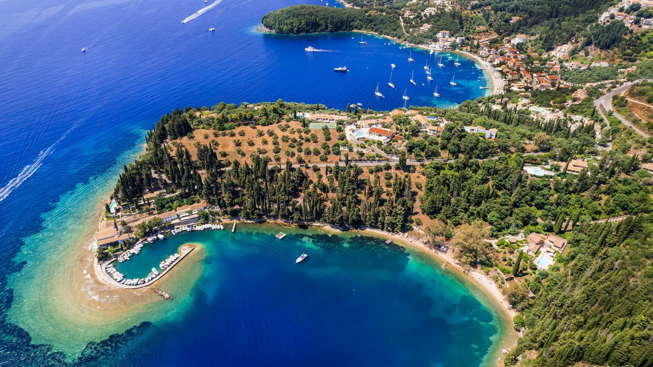 Aerial view of a coastal area with turquoise waters, a small marina with boats, a curved beach, and lush green trees and hills, along with a small seaside village.