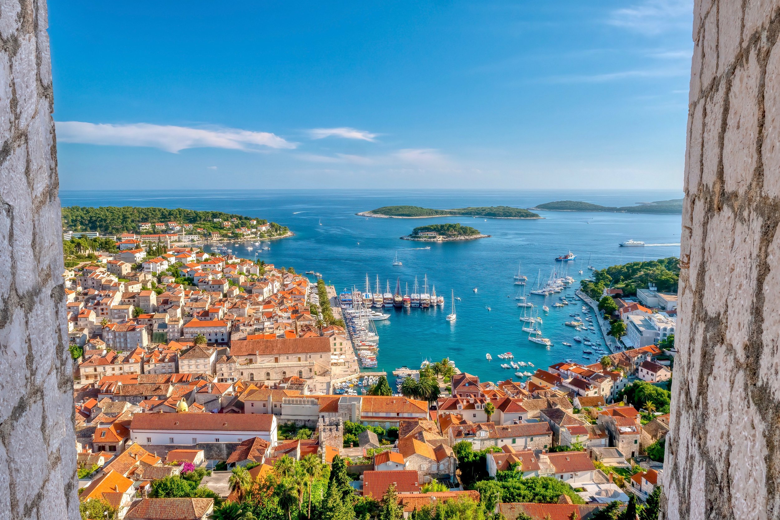 A scenic view of a small coastal town with red-roofed buildings overlooking a marina filled with sailboats, surrounded by lush green landscape and blue waters with islands and an ocean horizon.