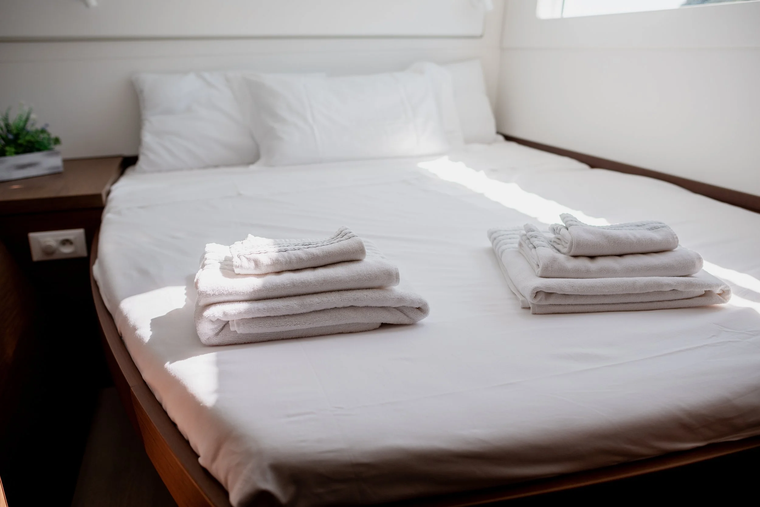 A neatly made bed with white linens and two stacks of folded white towels at the foot of the bed in a bright room with sunlight coming through a window.