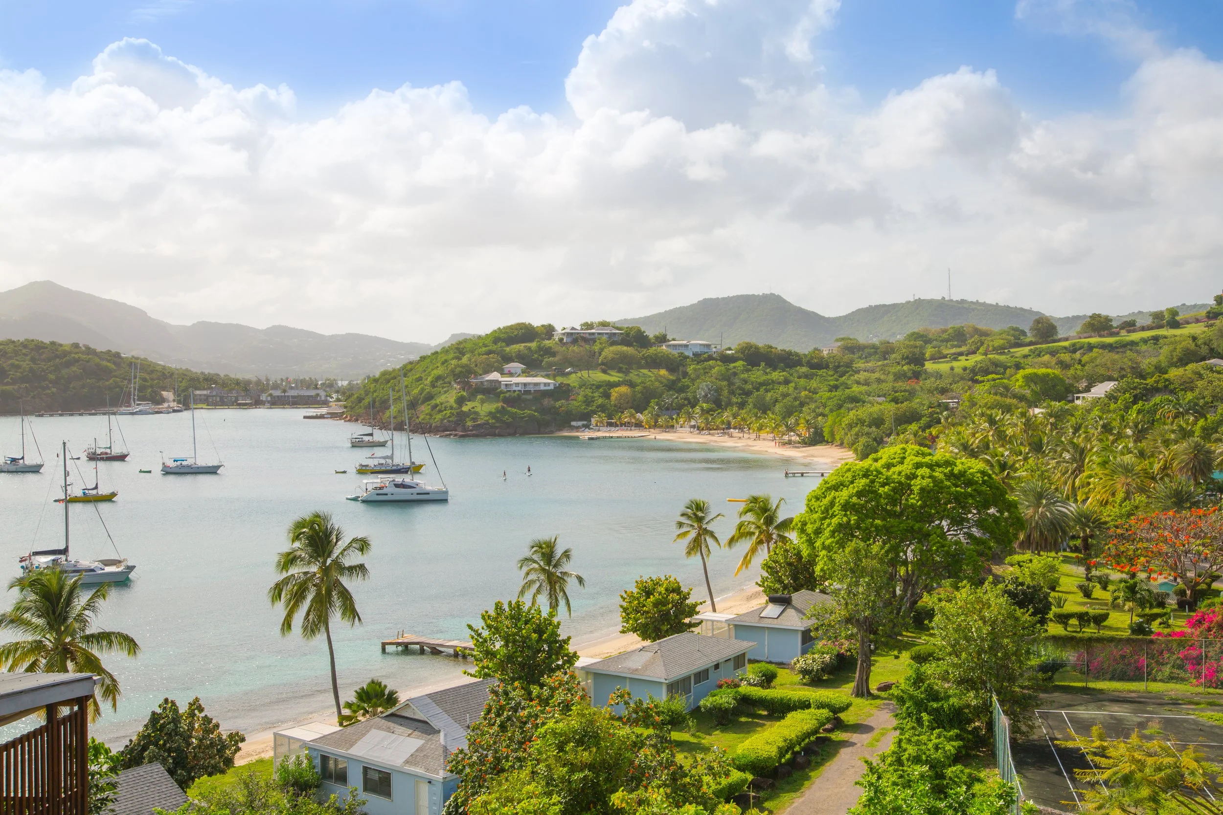 Scenic view of a tropical bay with sailboats anchored in calm water, lush green hills, palm trees, houses, and a sandy beach under partly cloudy skies.
