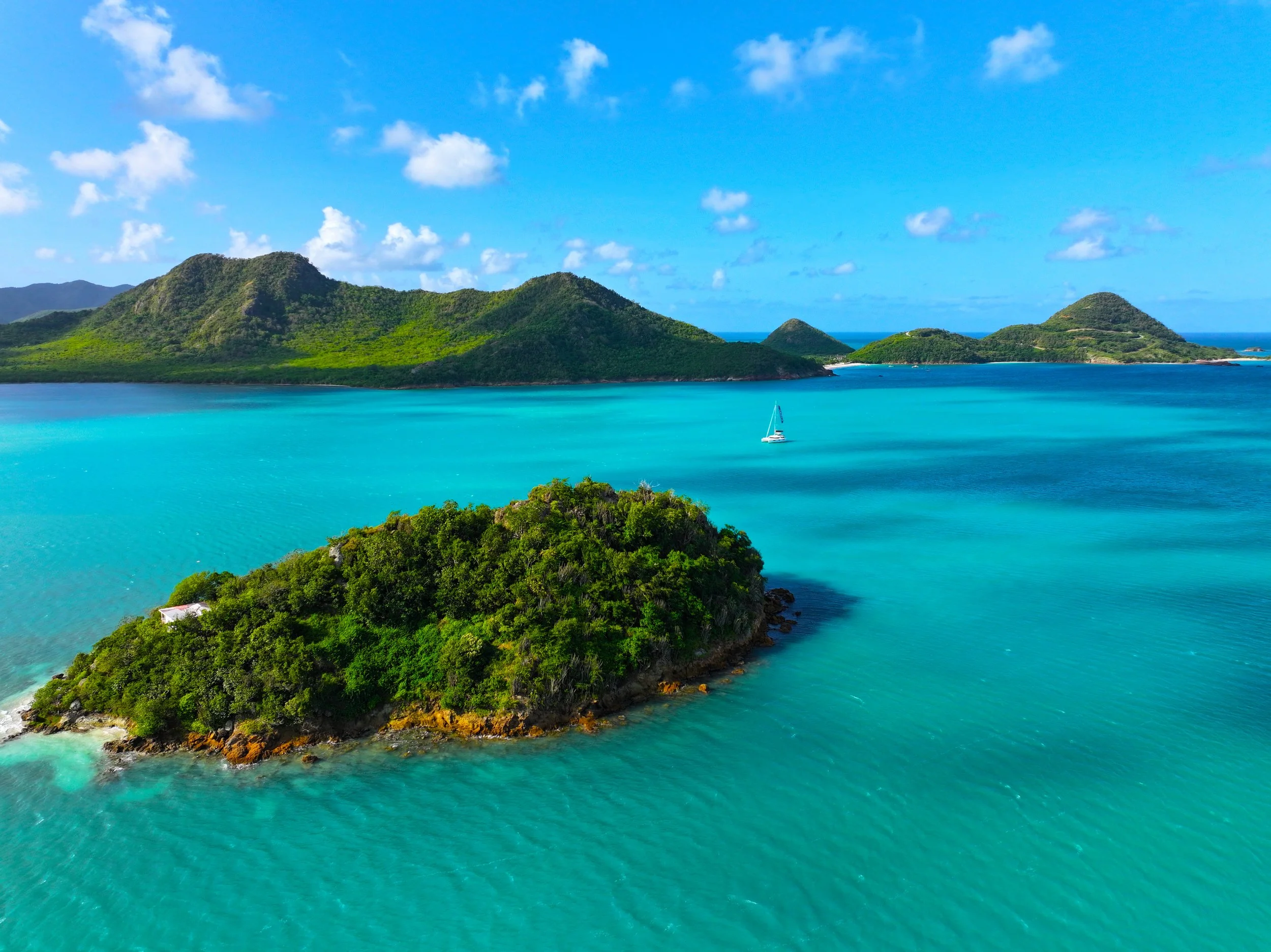 Tropical island with lush green vegetation surrounded by turquoise waters and a sailboat, with mountainous islands in the background under a partly cloudy sky.
