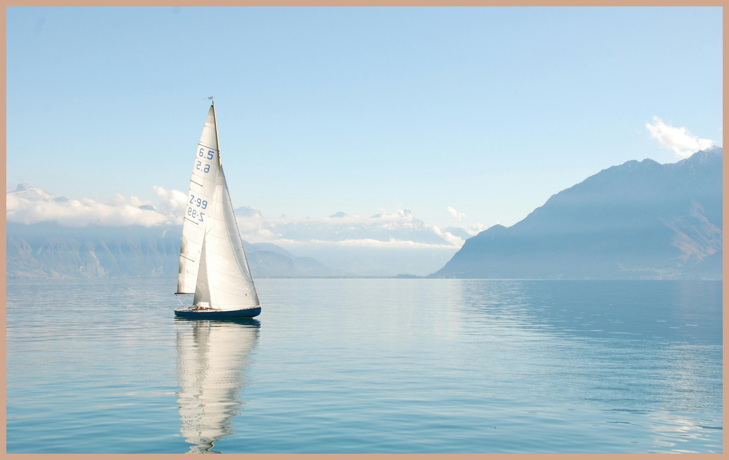 A sailboat on calm water with mountains in the background under a blue sky with scattered clouds.