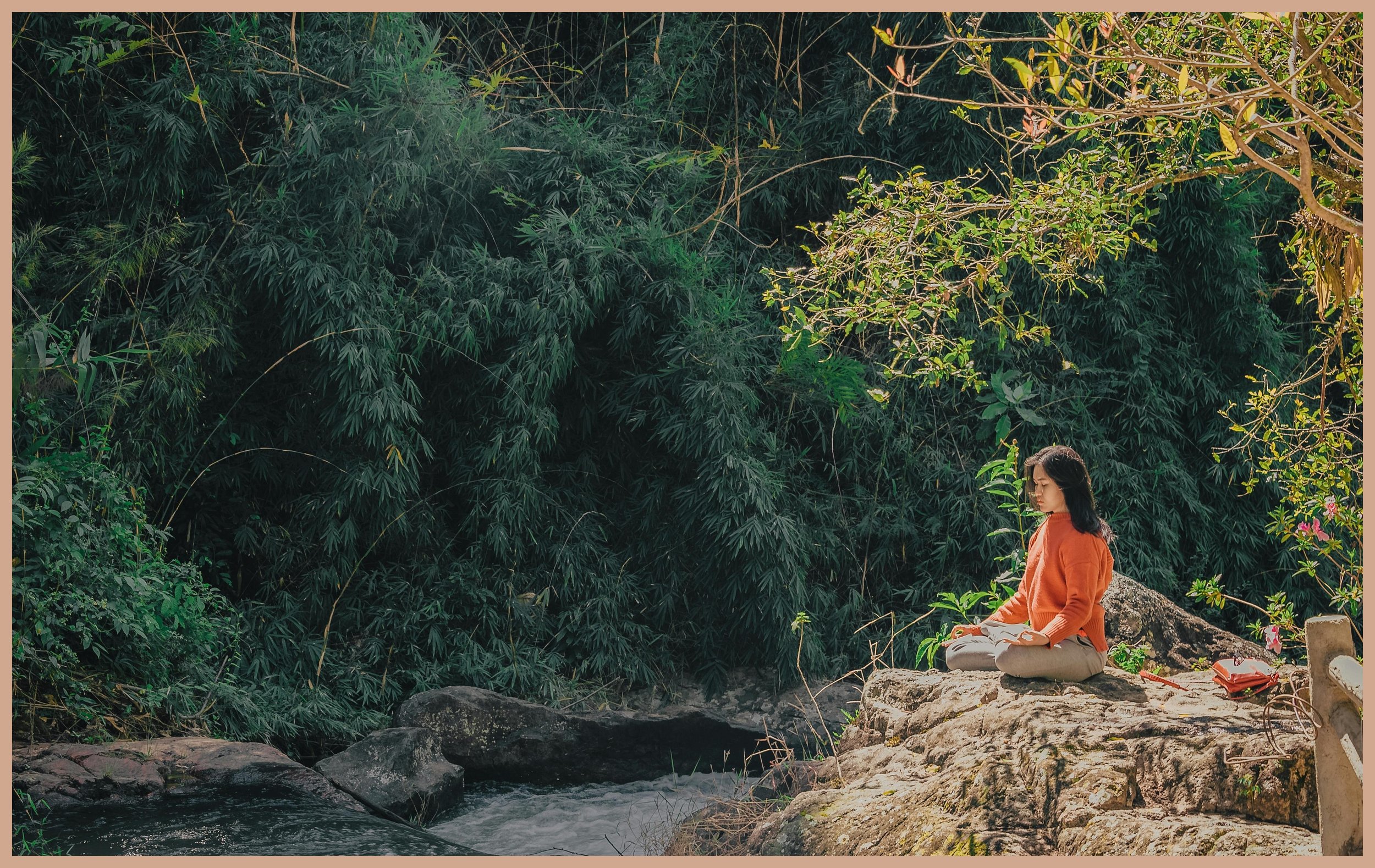 A woman sitting cross-legged on a rock by a stream in a lush green forest, practicing meditation.