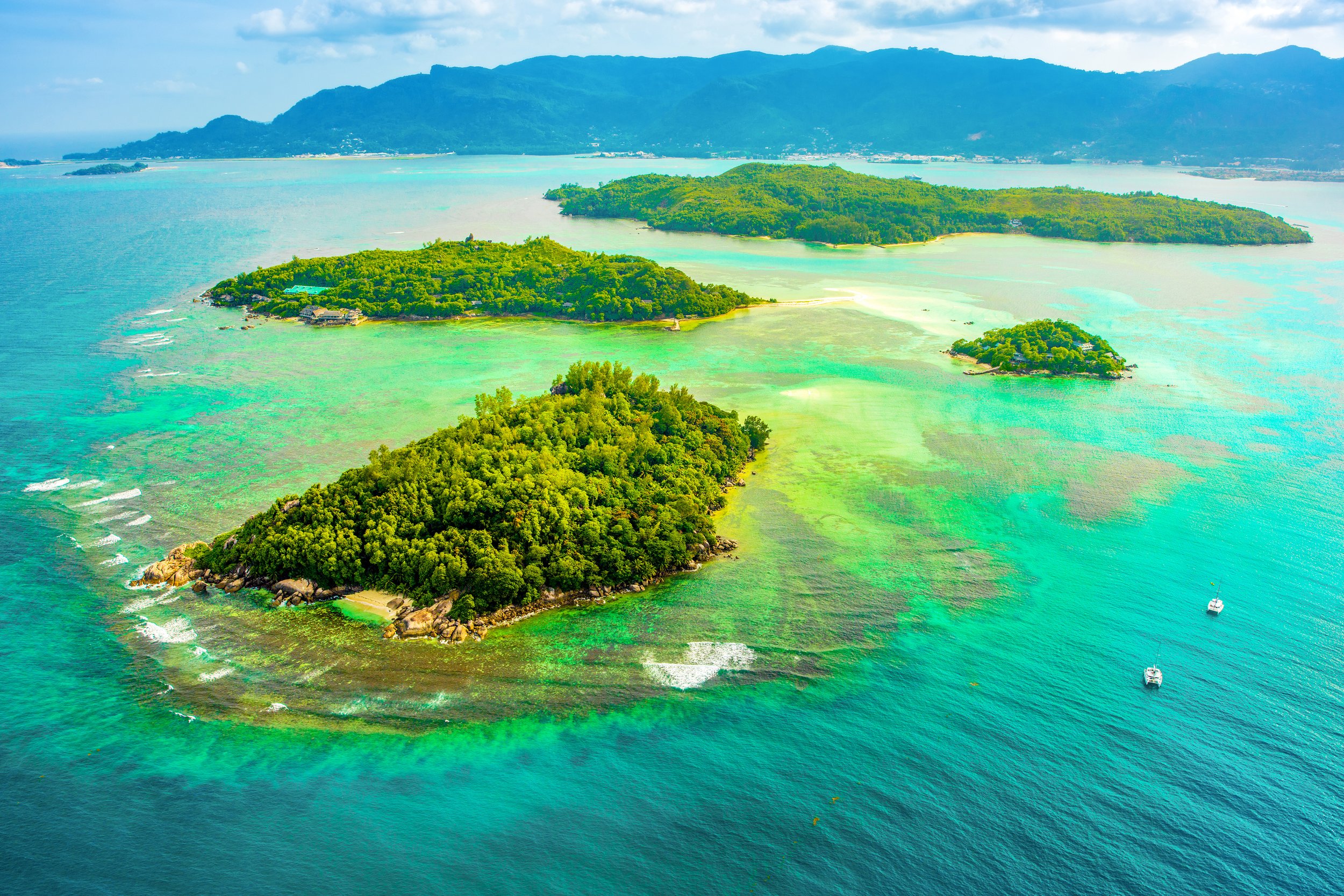 Aerial view of lush green islands surrounded by turquoise waters with boats in the water, mountains in the background.