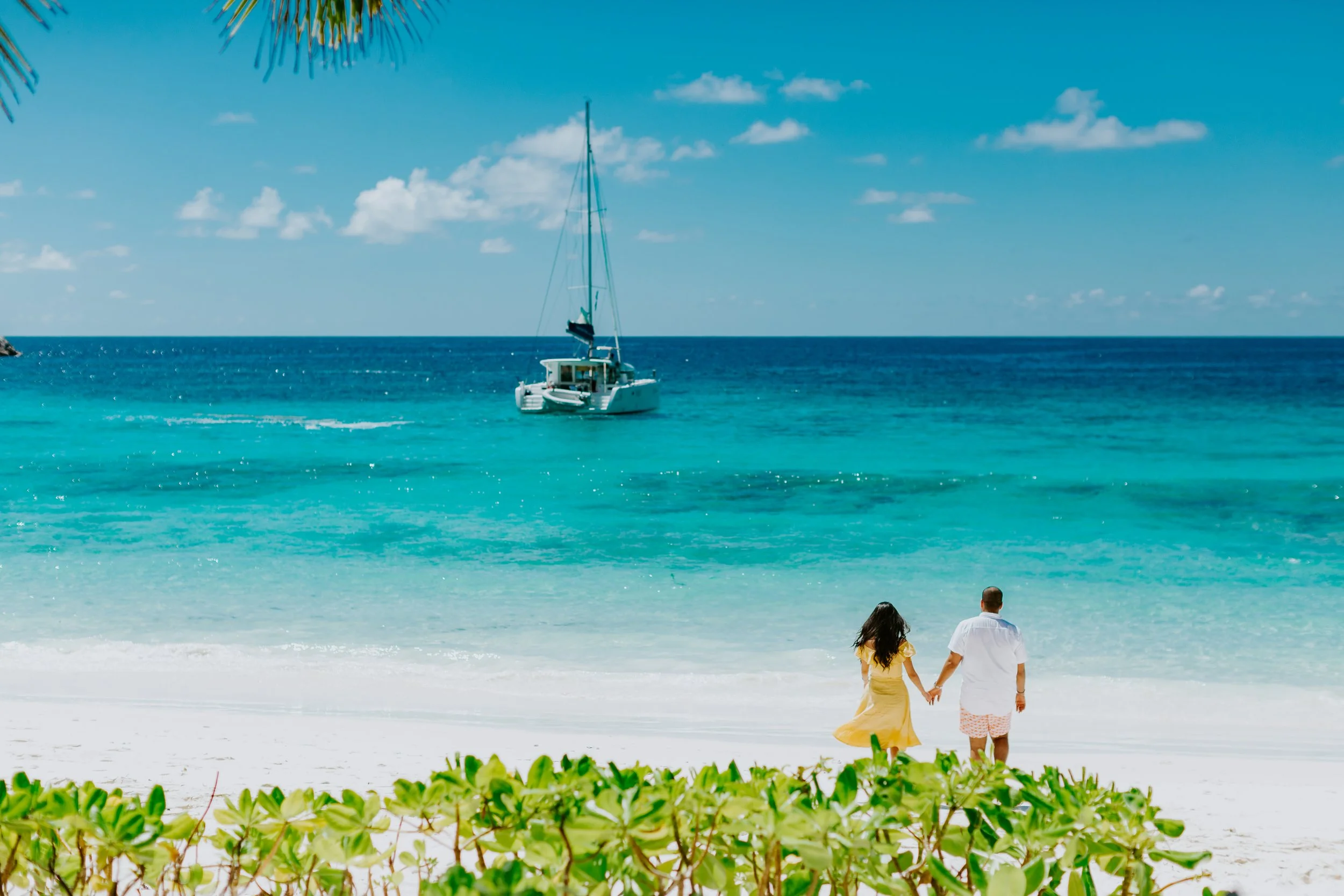 A couple holding hands and walking on the beach with clear turquoise water and a sailboat in the distance under a partly cloudy sky.