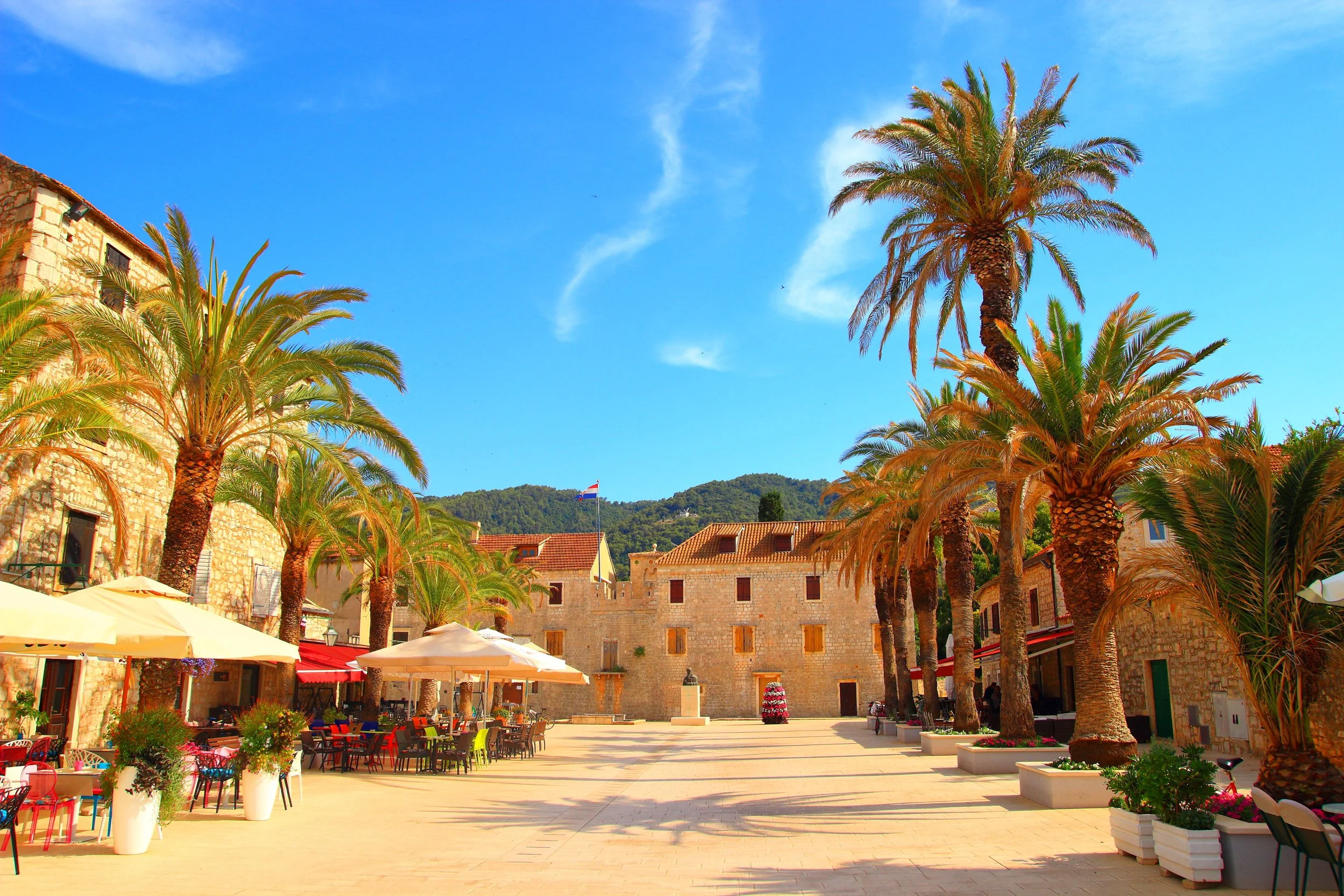 A sunny outdoor plaza with palm trees, outdoor seating with umbrellas, and stone buildings, in a Mediterranean-like setting.