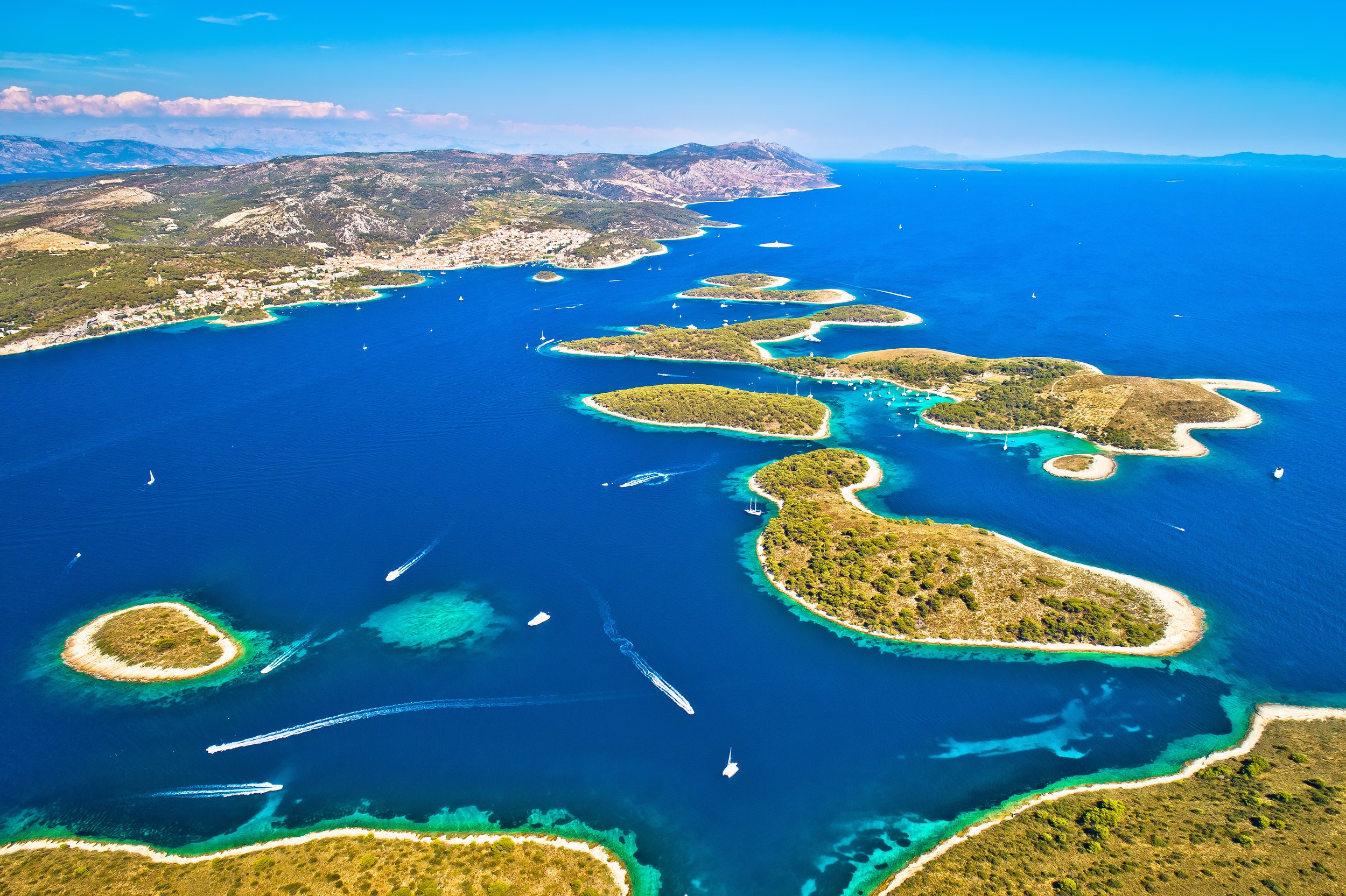 Aerial view of a coastline with small islands, clear blue water, and boats sailing in a harbor, with a mountainous landscape in the background.