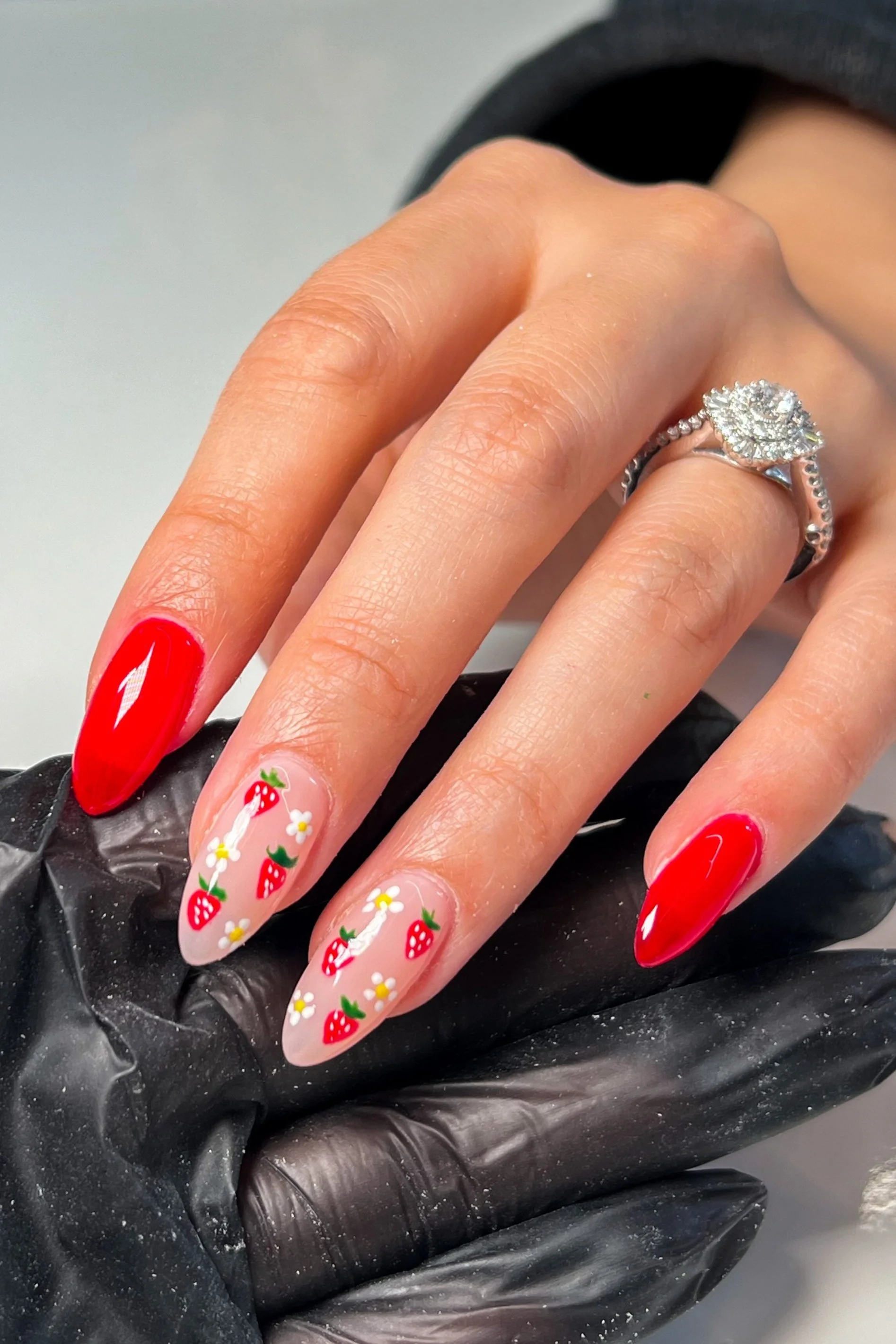 Nail art with red lacquer on the thumb and pinky, and strawberries with white flowers on a clear base on the middle and ring fingers, a silver ring with a large, round, diamond-like gemstone on the ring finger.