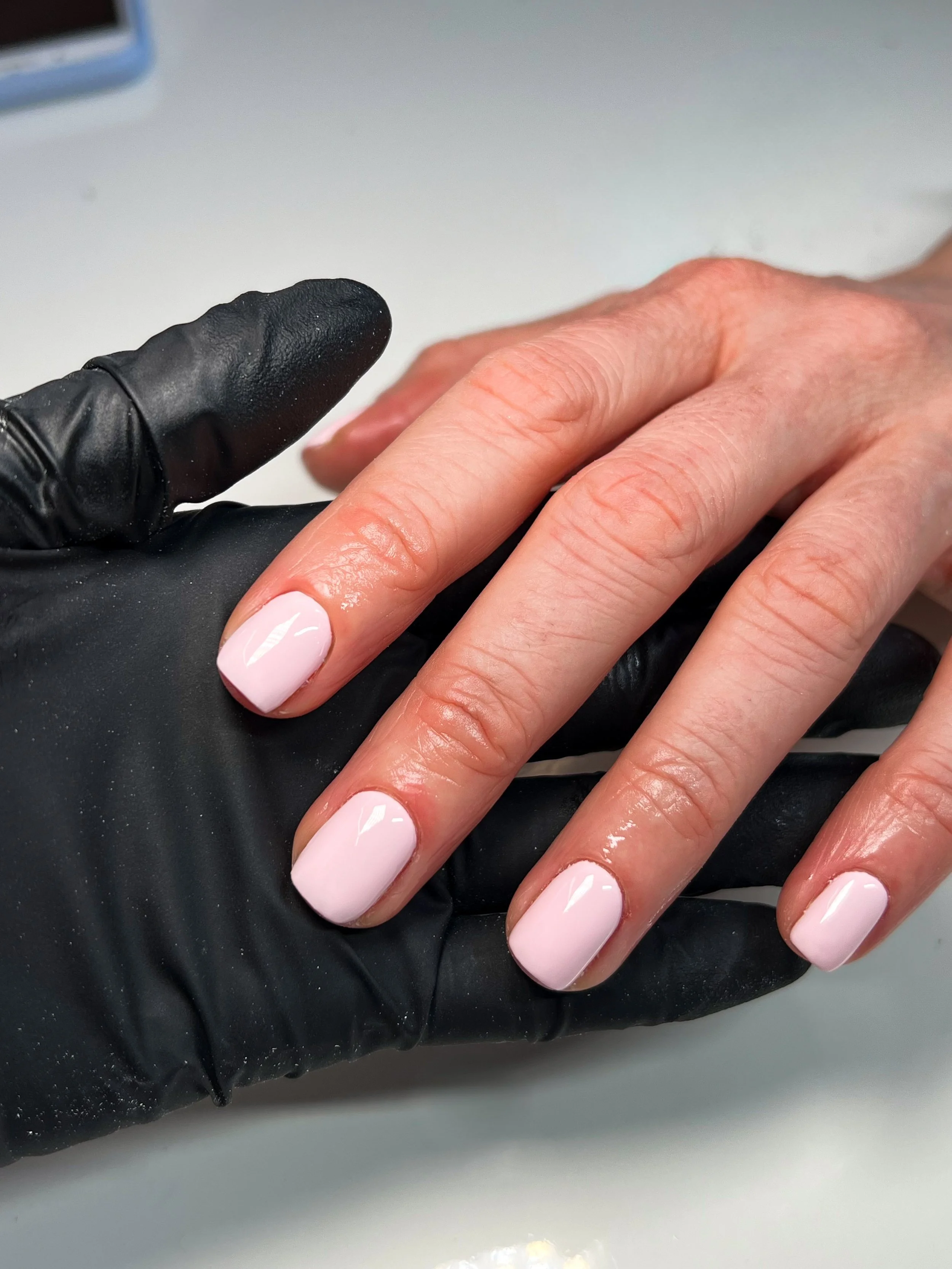 A person with well-manicured shellac light pink nails getting a manicure, wearing a black glove on the hand holding the nails.