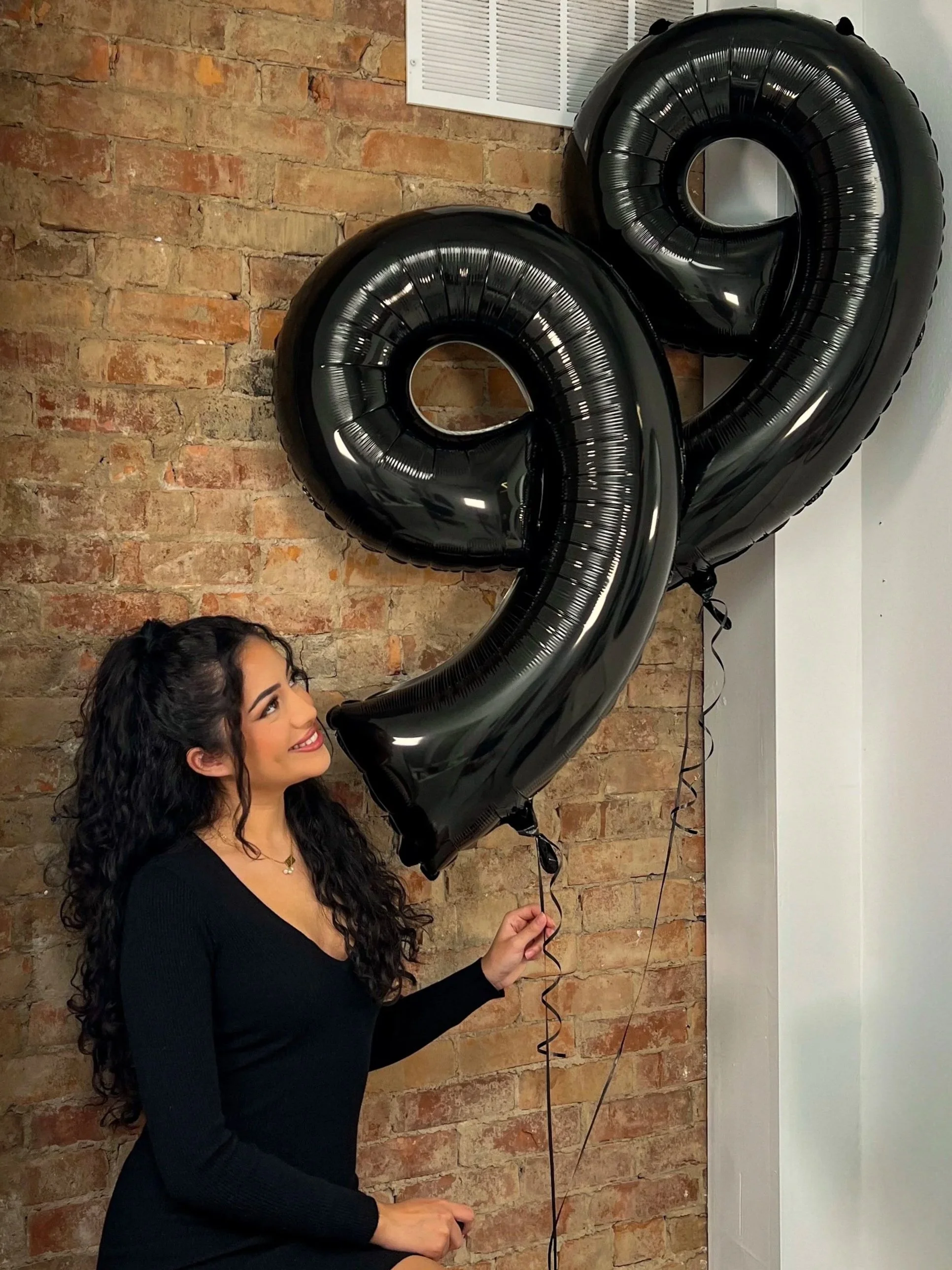 A woman with long curly hair, wearing a black dress, is holding black number balloons in the shape of 9 and 2, standing against a brick wall for a celebration.
