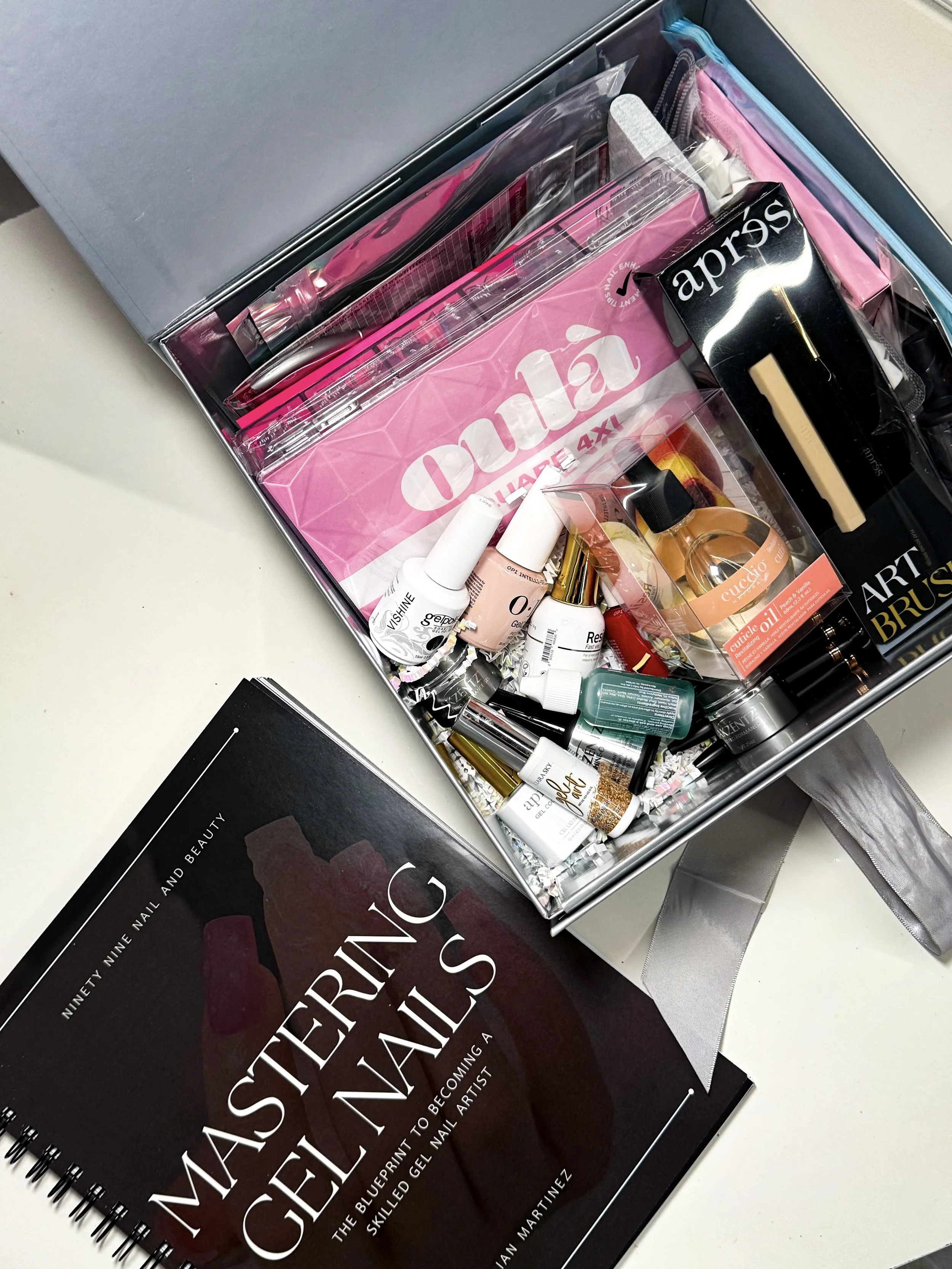 A round metal tin open to reveal various nail art supplies, including nail polish bottles, brushes, and nail files. Beside it, a book titled 'Mastering Gel Nails'.