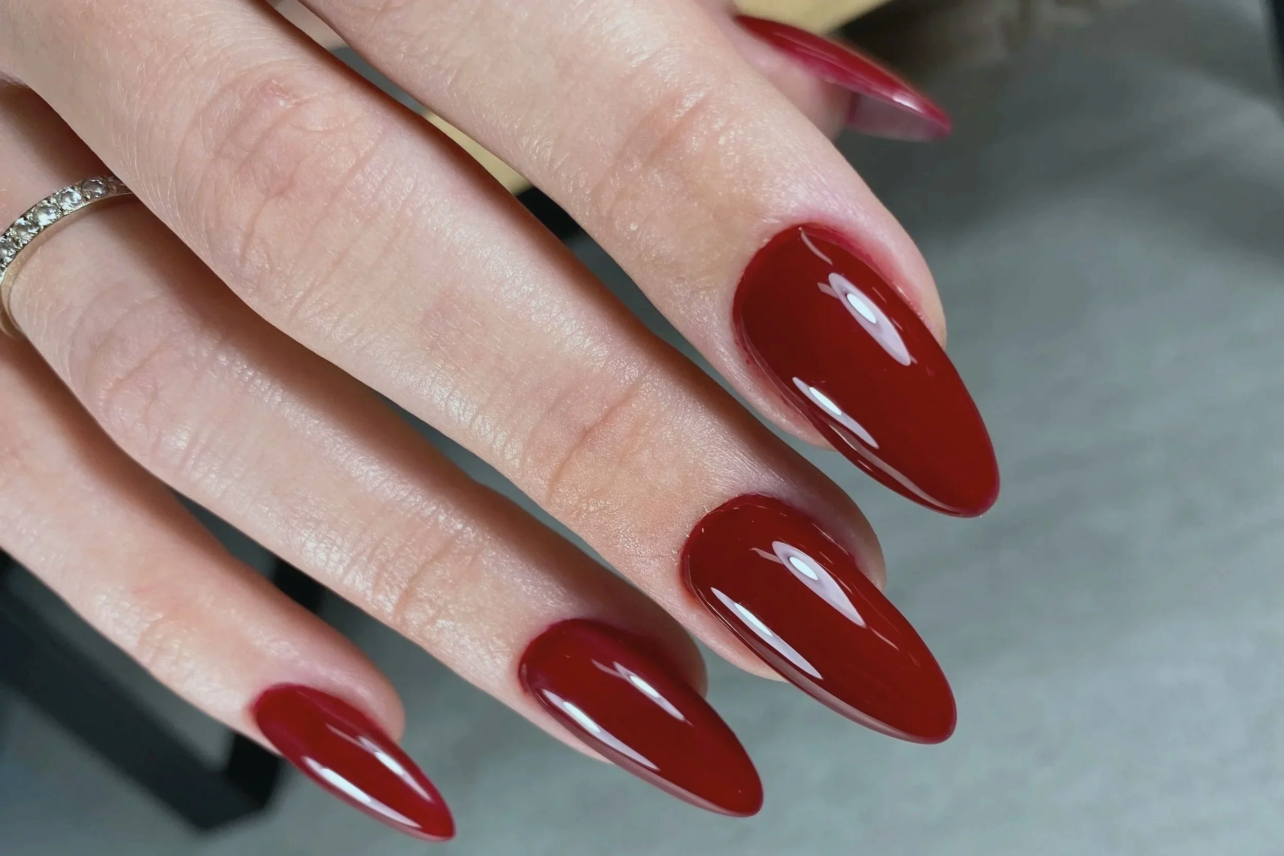 Close-up of a hand with long, almond-shaped nails painted in shiny dark red nail polish, and a silver ring with small gemstones on the ring finger.