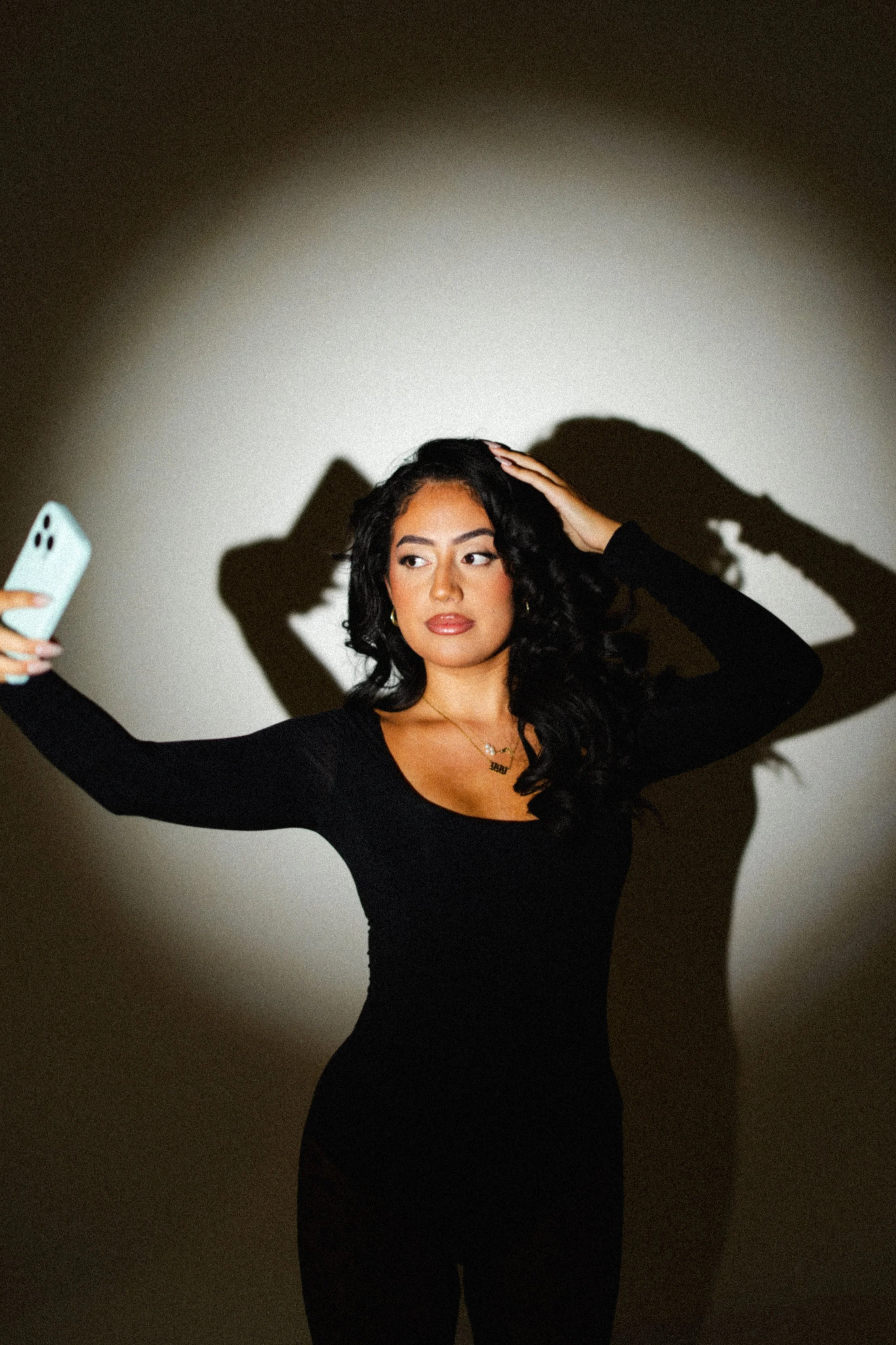 A woman taking a selfie with a smartphone, standing against a plain wall with a shadow cast behind her.