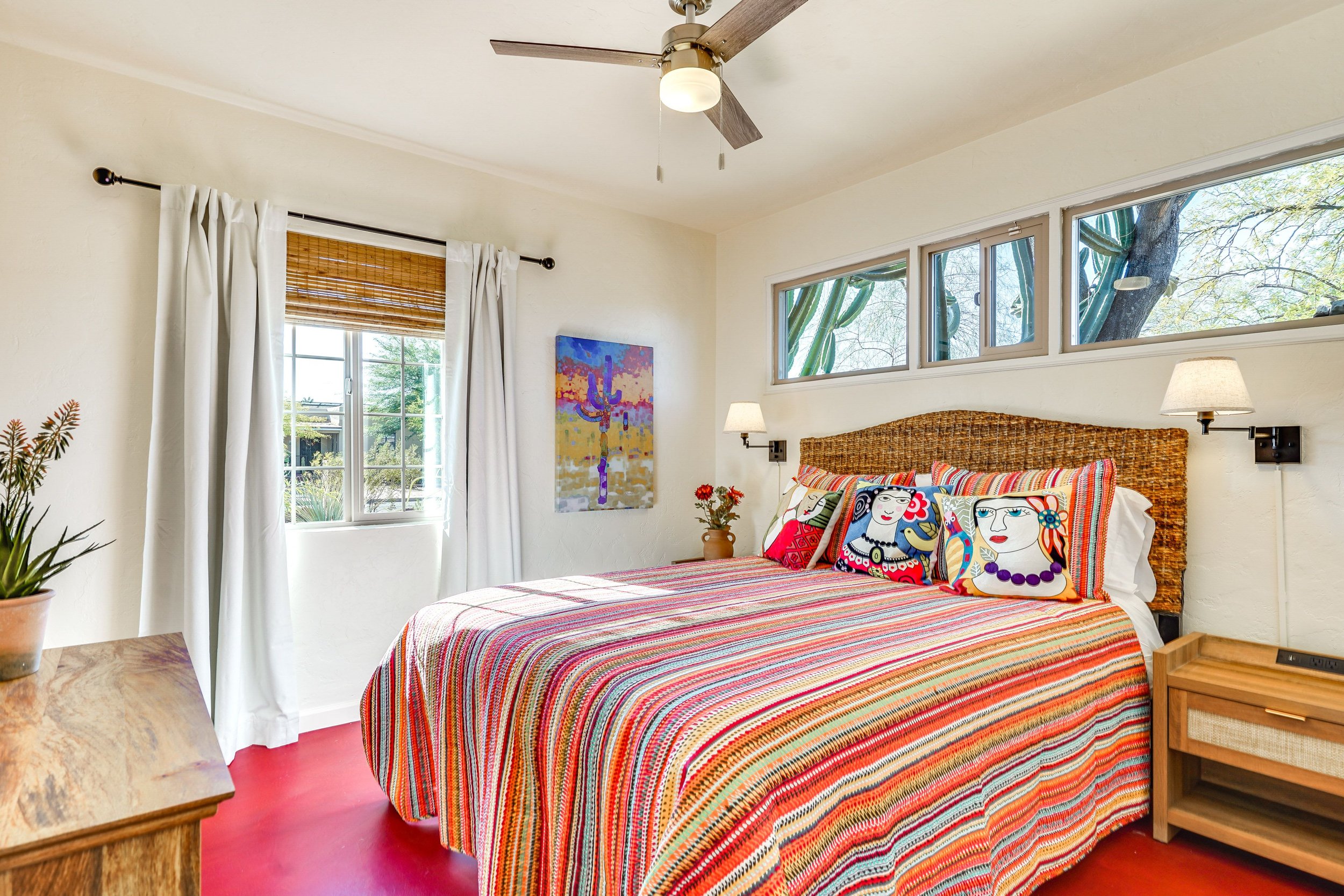A bright bedroom with a colorful striped bedspread, vibrant pillows featuring abstract designs, wicker headboard, ceiling fan, wall art, potted plant, wooden side tables, and large windows with outside view.