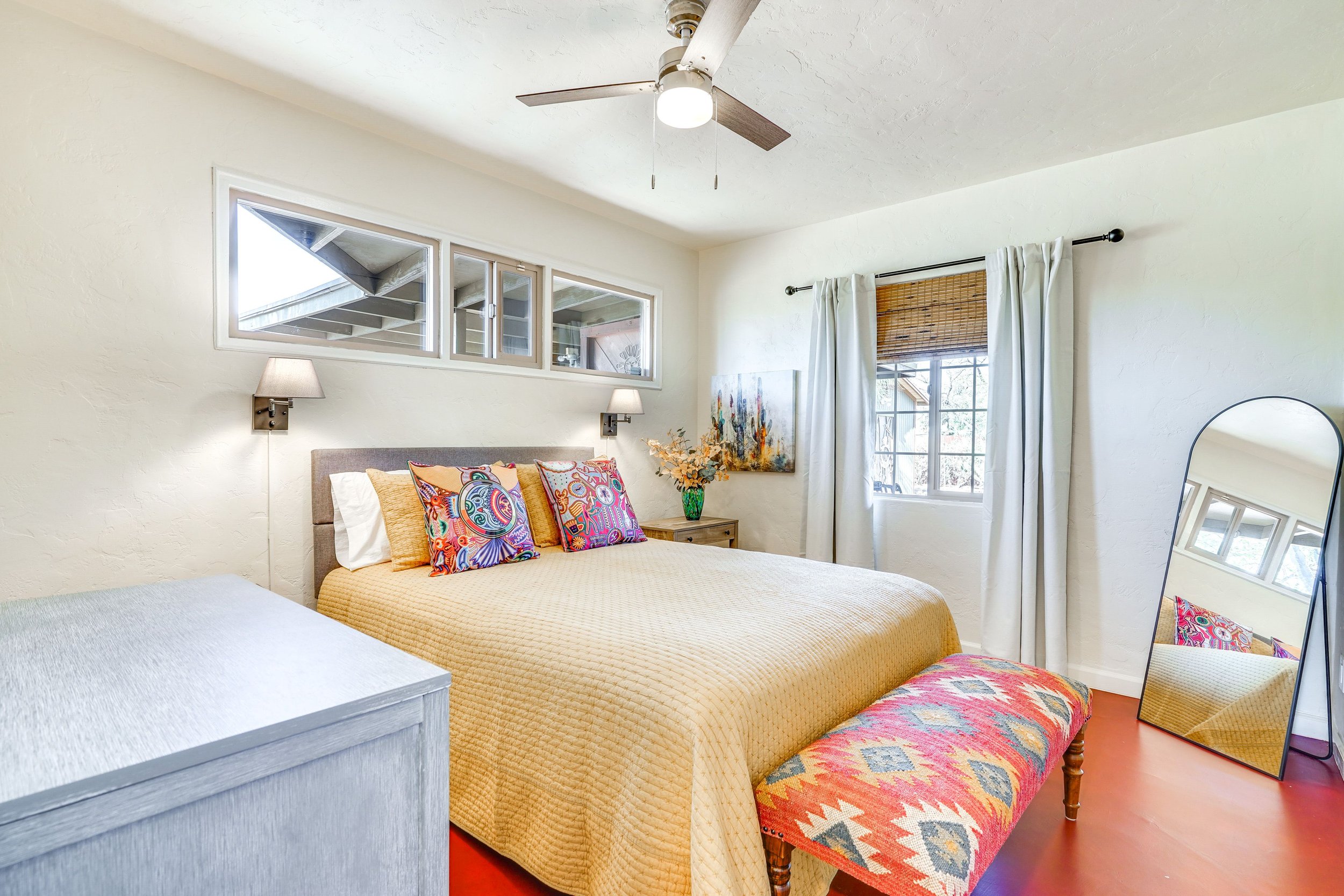 Bright bedroom with a beige bedspread, colorful patterned pillows, wall-mounted lamps, ceiling fan, curvy full-length mirror, wooden nightstand with flowers, rectangular window with light curtains, and a vibrant patterned bench at the foot of the bed