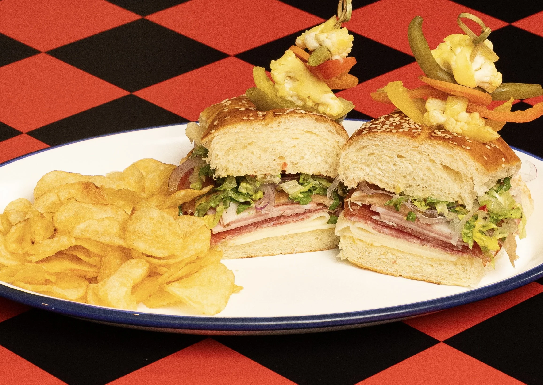 Sub sandwich with meats, cheese, lettuce, and vegetables, served with potato chips and topped with pickled vegetables on a white plate.