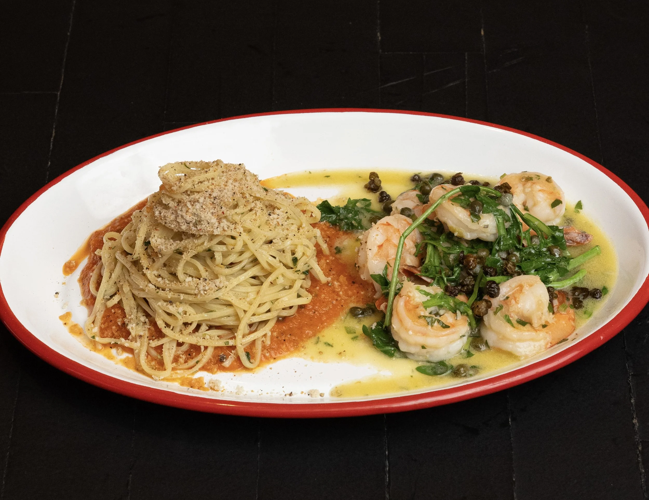 A white oval plate with a red rim holding a serving of spaghetti in tomato sauce topped with grated cheese on the left and sautéed shrimp with a green herb sauce and black peppercorns on the right.