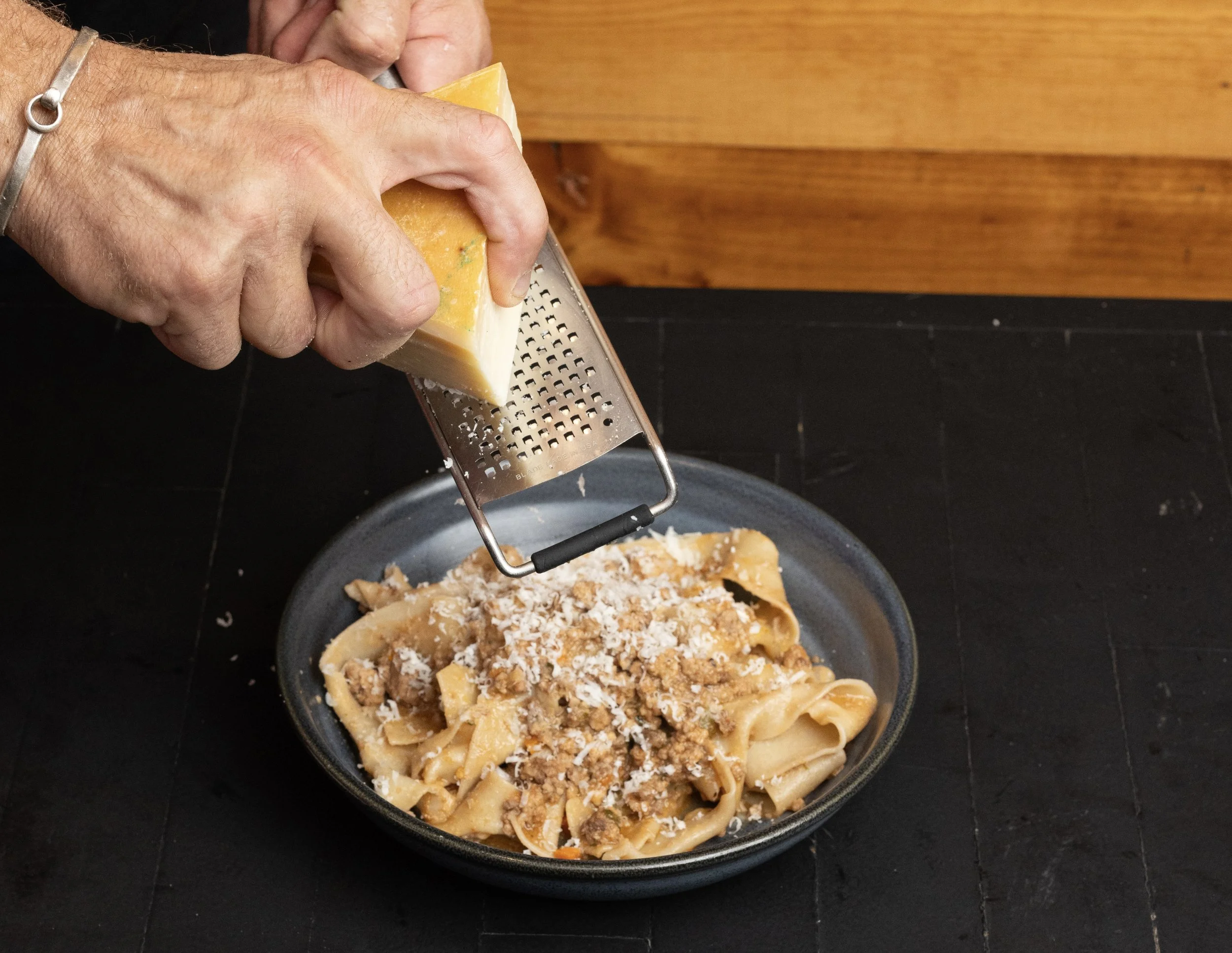 Close-up of hands grating cheese over a bowl of cooked pasta with meat sauce