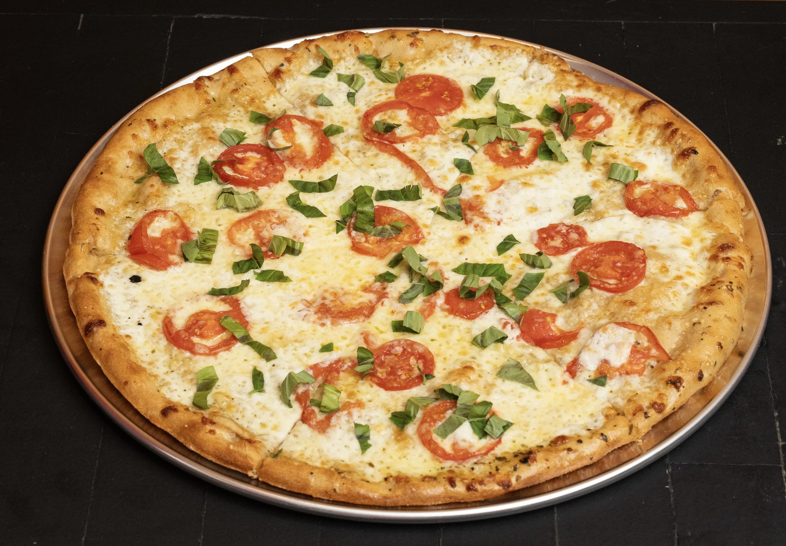 A cheese pizza topped with sliced tomatoes and chopped basil, on a round tray.