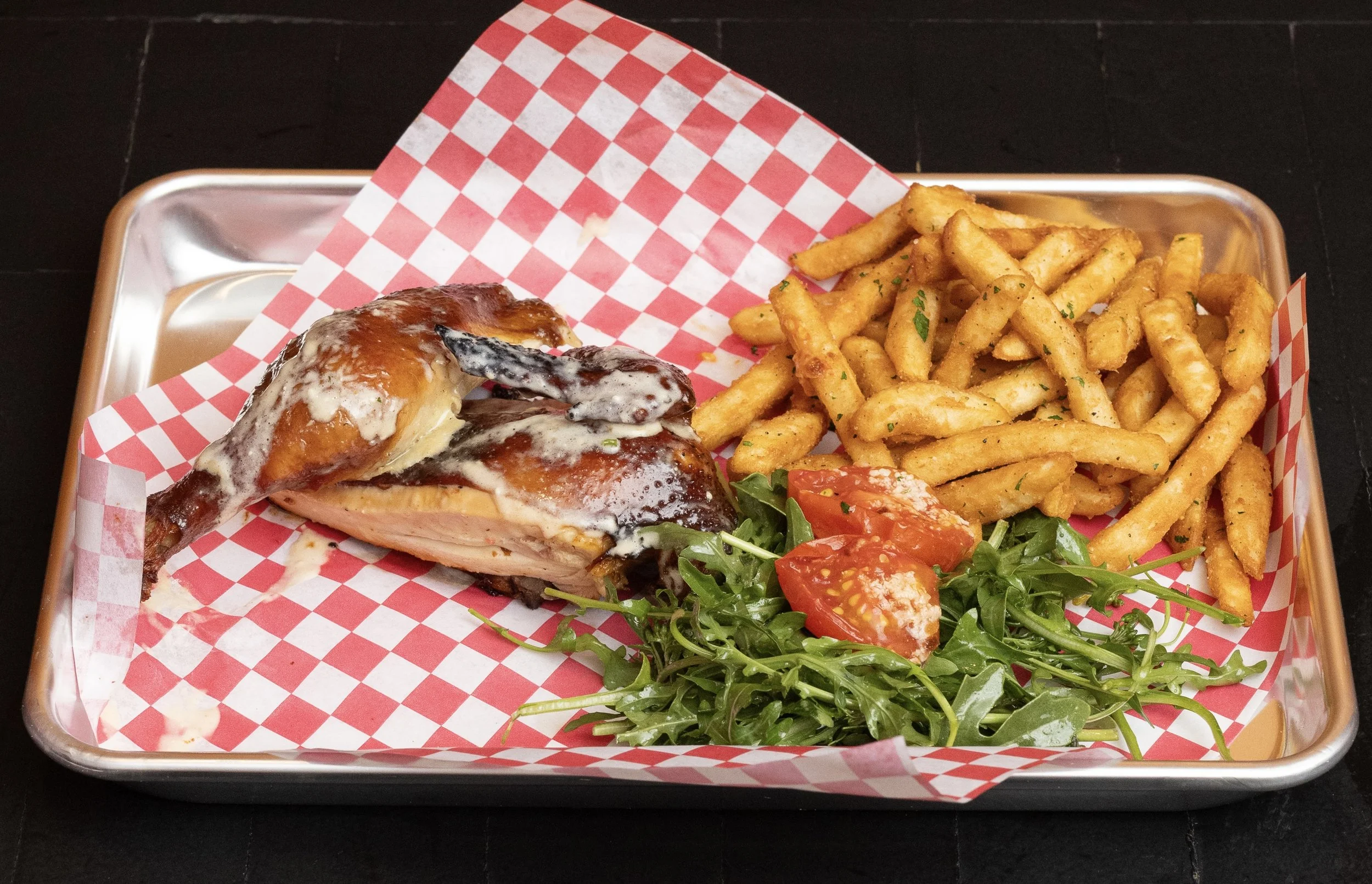 Tray of food with roasted chicken leg, French fries, tomato cucumber salad, and arugula garnished with herbs on red and white checkered paper