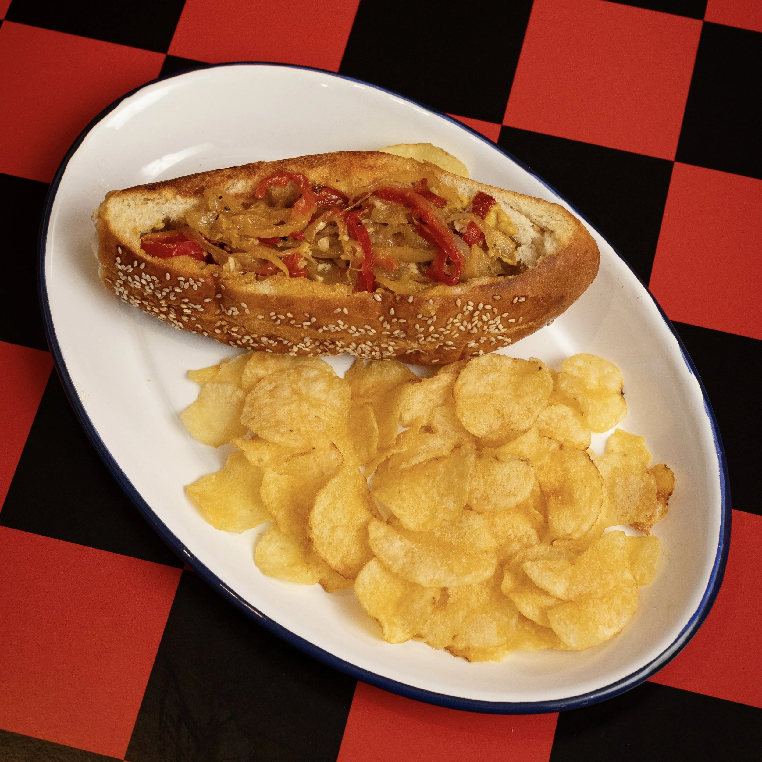 A white plate with a sesame seed-covered hot dog in a bun topped with chili and chopped peppers, and a generous serving of potato chips, set on a black and red checkered table.