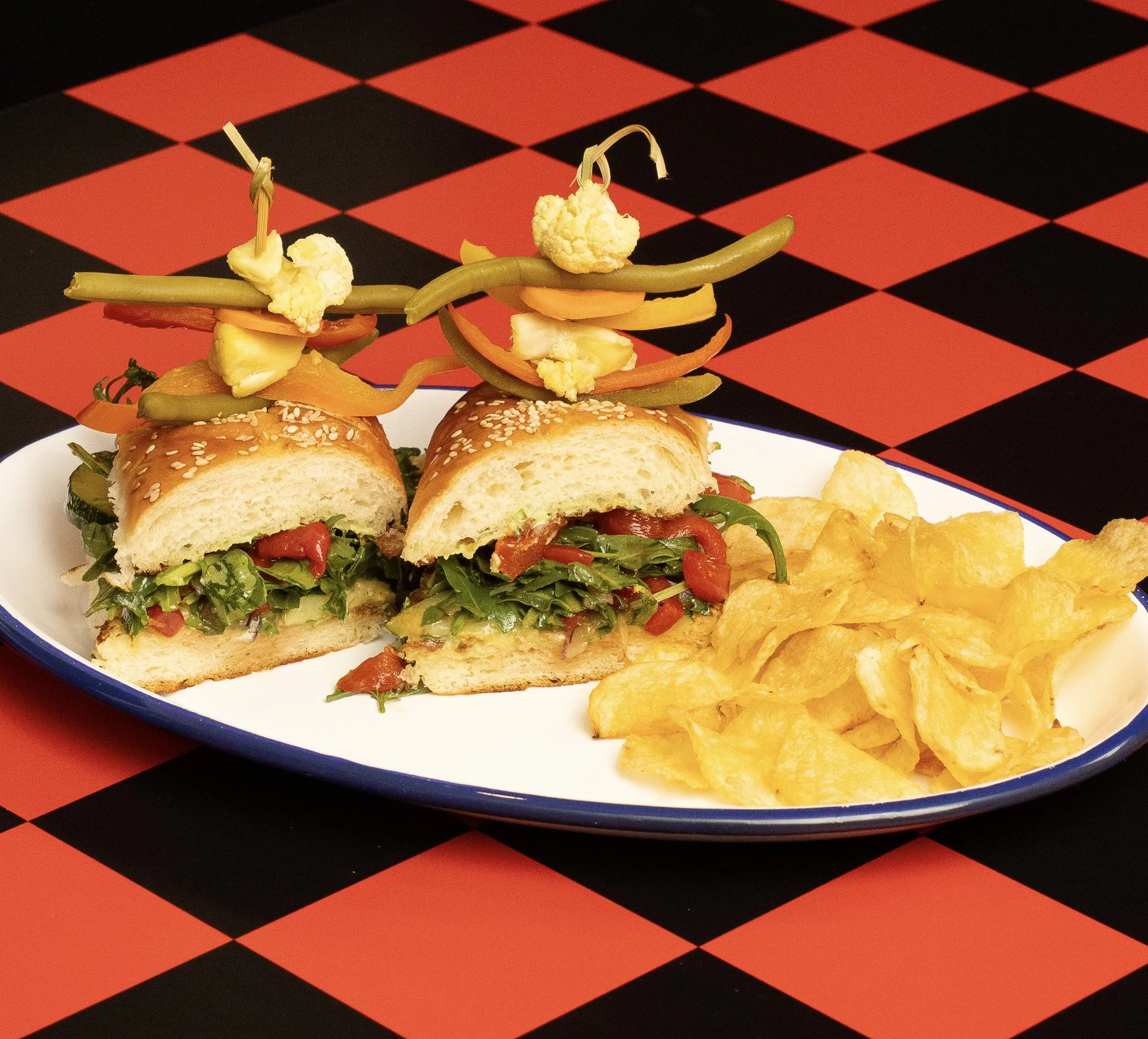 Sandwiches with lettuce, tomato, and green peppers, with decorative pickles and cauliflower on top, served with potato chips on a white plate, on a red and black checkered tablecloth.