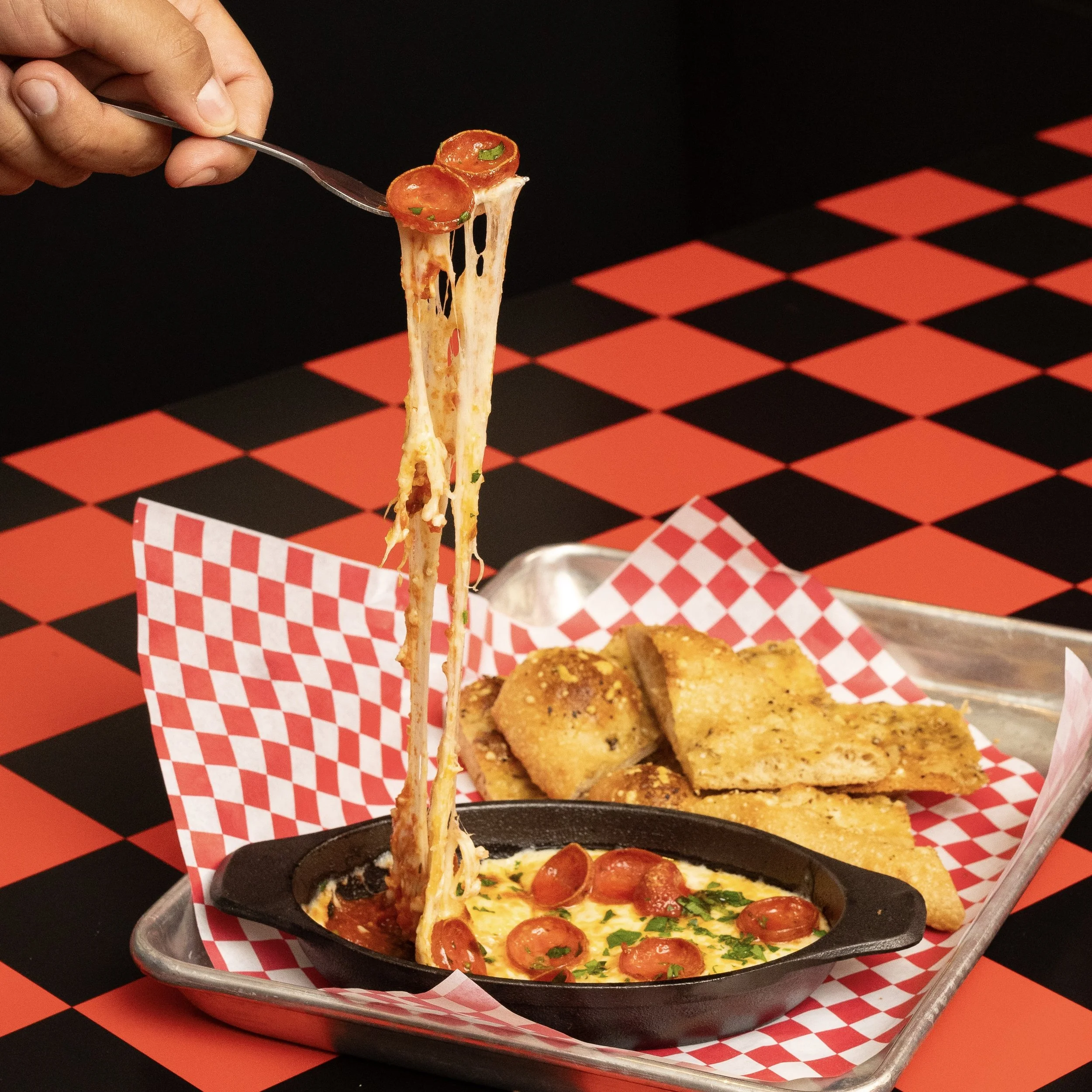 A hand is lifting a slice of pepperoni pizza with melted cheese; the pizza is on a black skillet on a metal tray lined with red and white checkered paper, along with garlic bread sticks on a red and black checkered table.