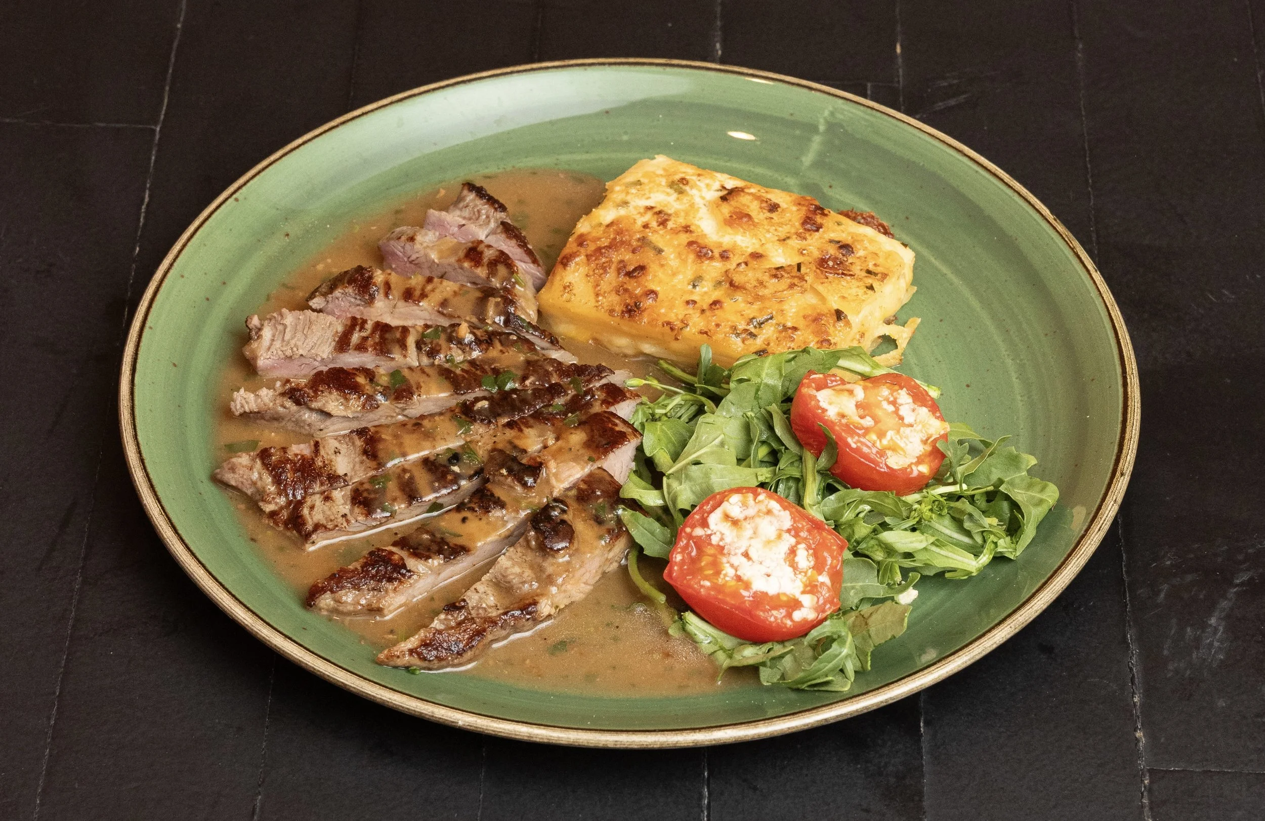 A green oval plate with sliced grilled steak, a baked potato, and a small salad with cherry tomatoes and cheese.