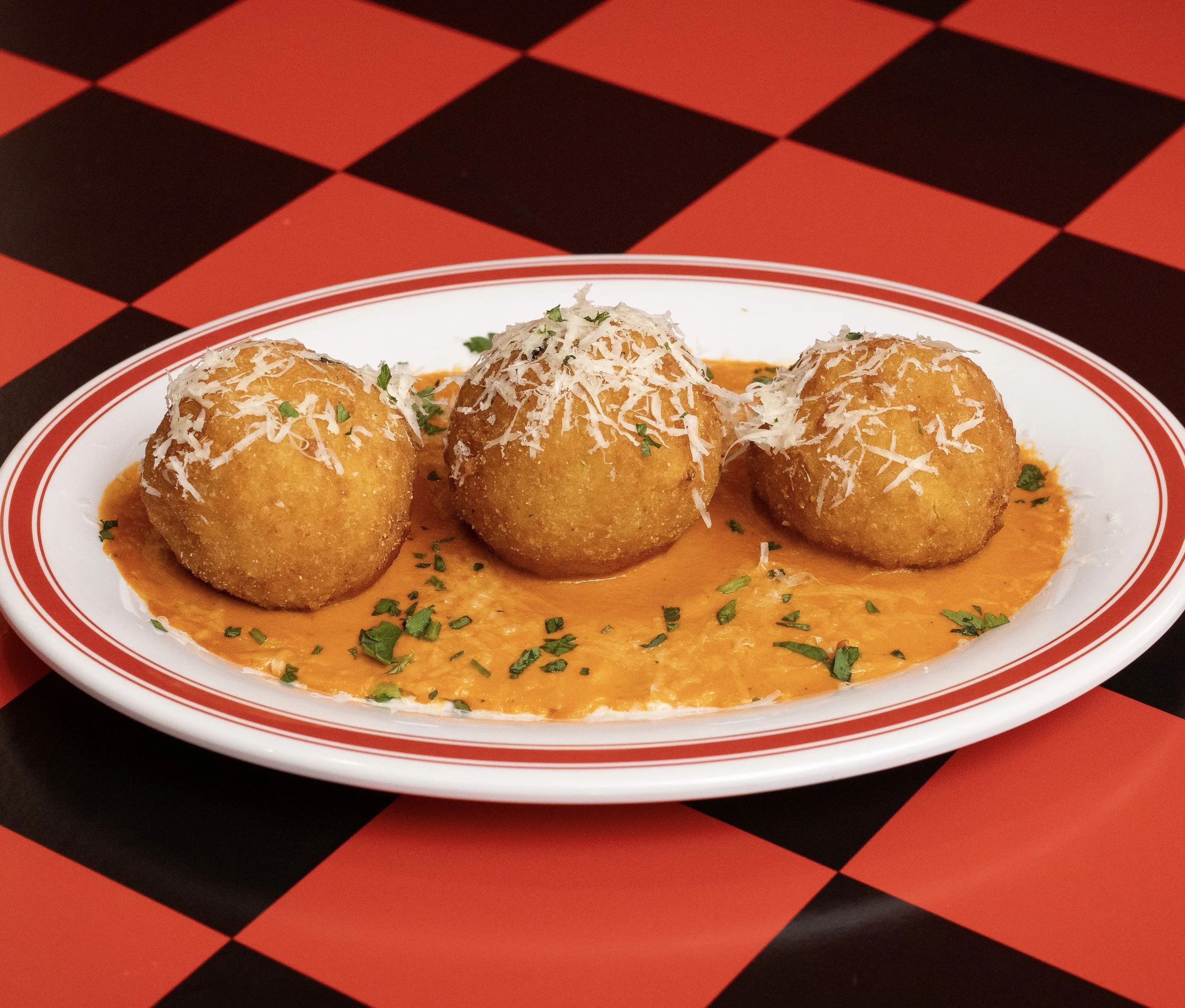 Three fried cheese balls topped with shredded cheese and chopped herbs, served on a white plate with orange sauce, on a red and black checkered table.