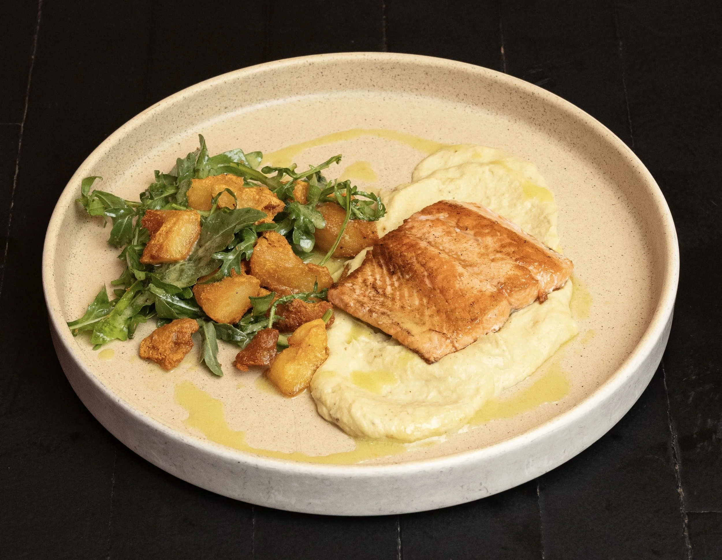 A plate with cooked salmon fillet on a bed of mashed potatoes, garnished with olive oil, next to a side salad with greens and croutons.