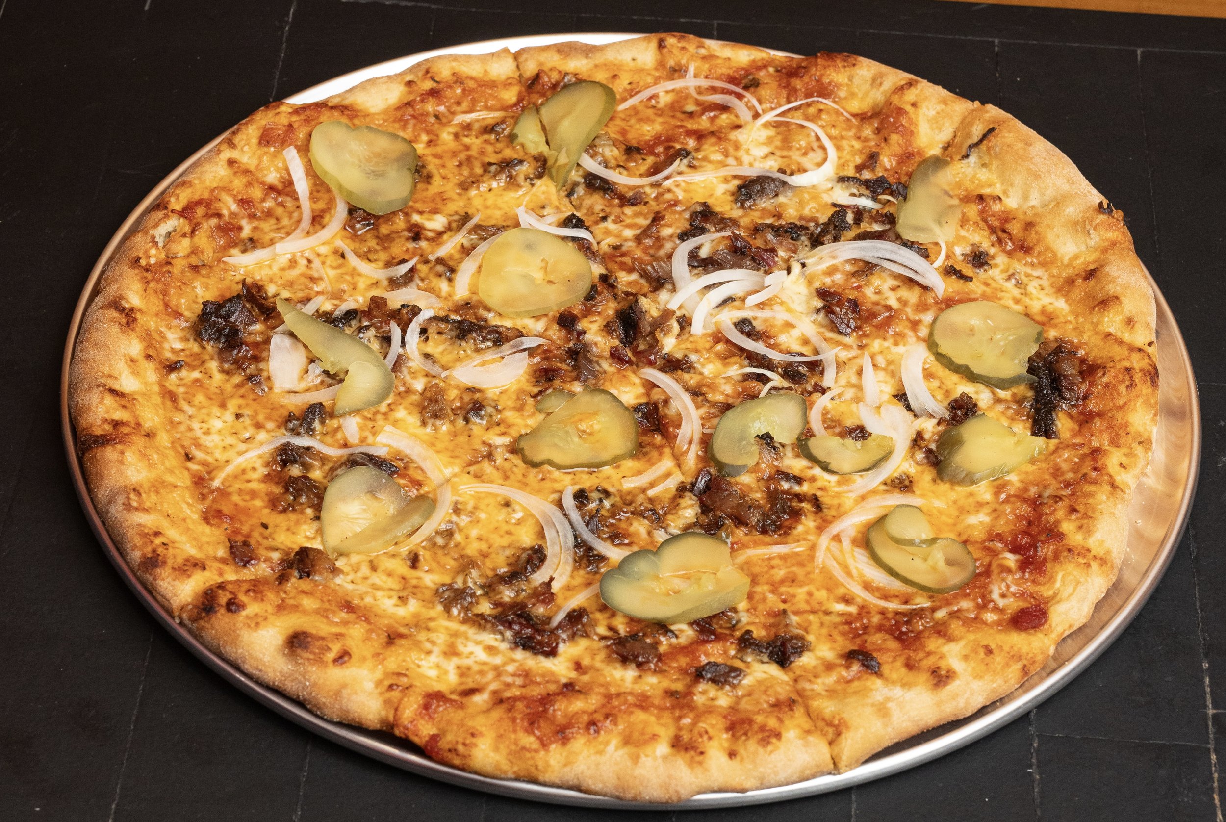 A whole pizza with cheese, pickles, onions, and bacon on a round metal tray.