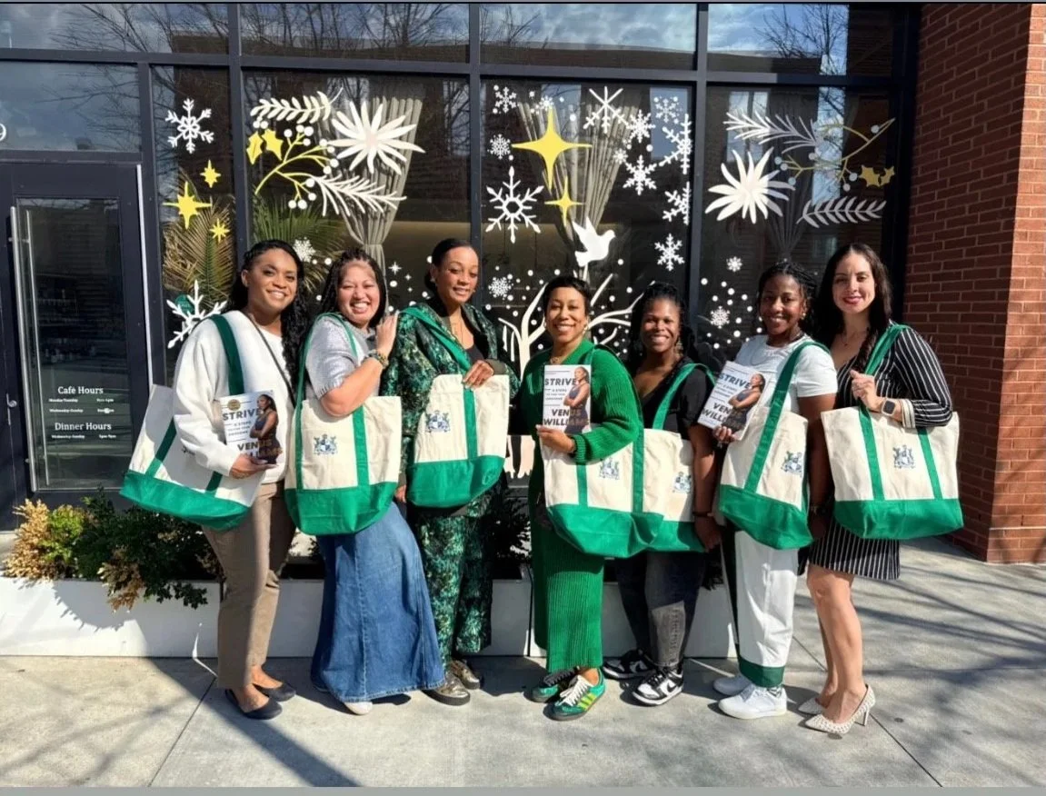 Group of seven women standing outside a glass storefront with holiday decorations on the window. They are holding tote bags and copies of a book titled "STRIVE." The women are smiling and dressed casually and professionally.