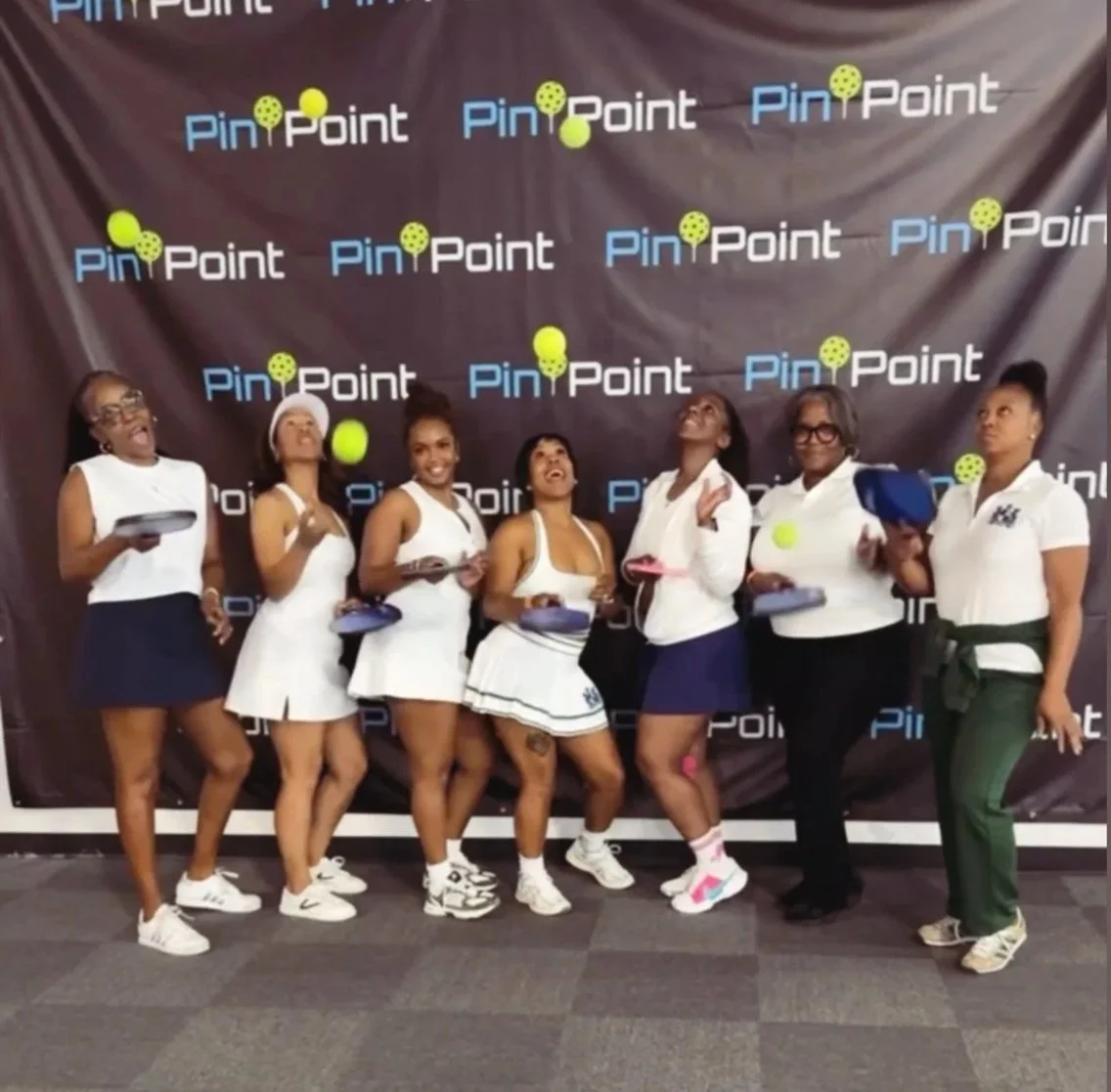 Group of women dressed in tennis outfits standing in front of a black backdrop with 'Pin Point' logo, some holding tennis balls and paddles, smiling and posing for a photo.