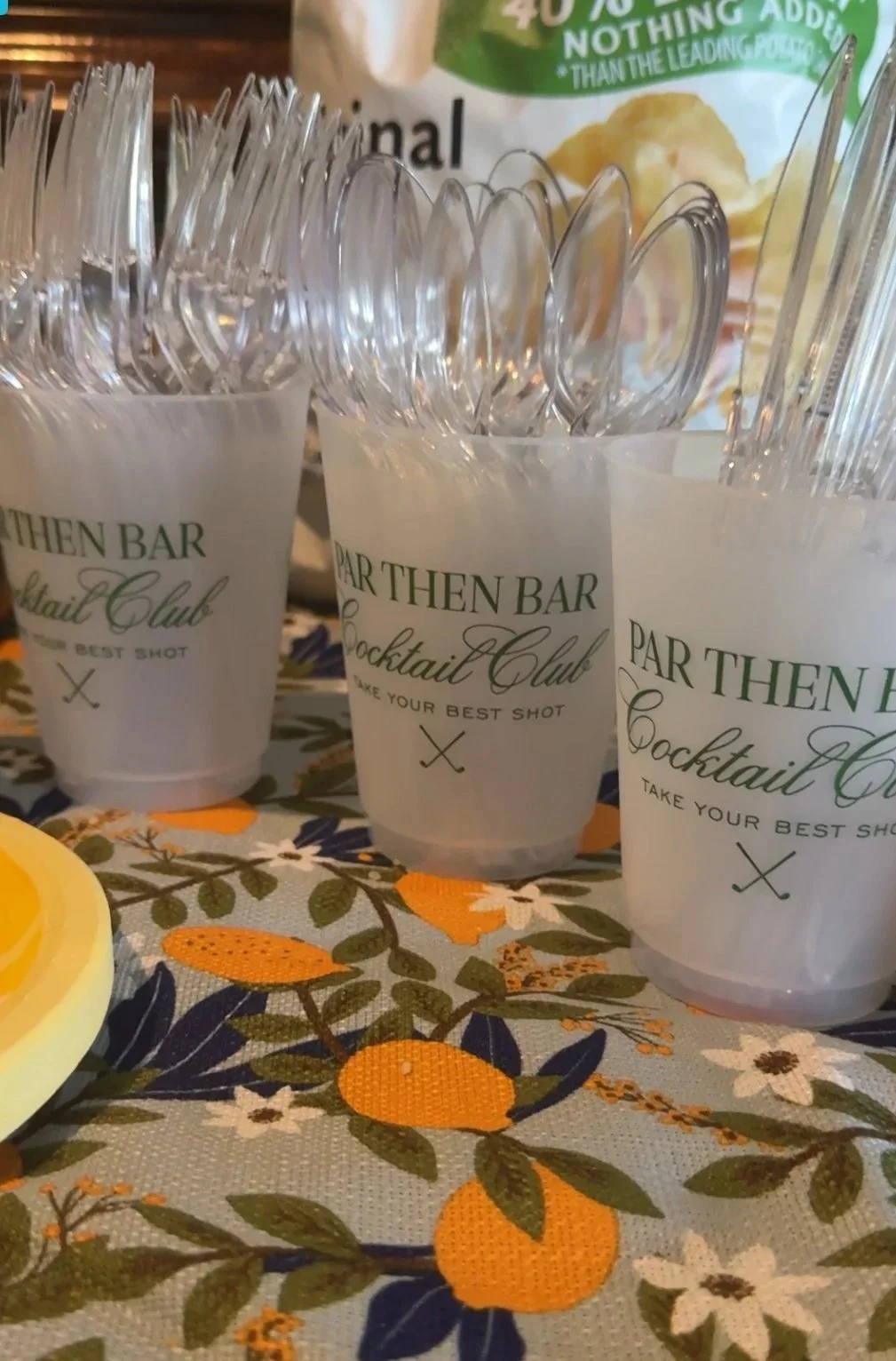 Clear plastic cups with green text reading 'Parthenon Bar Cocktail Club' and 'Take your best shot' are filled with utensils and placed on a floral tablecloth with orange, white, and green patterns, with a partially visible lemon and a bag of potato c