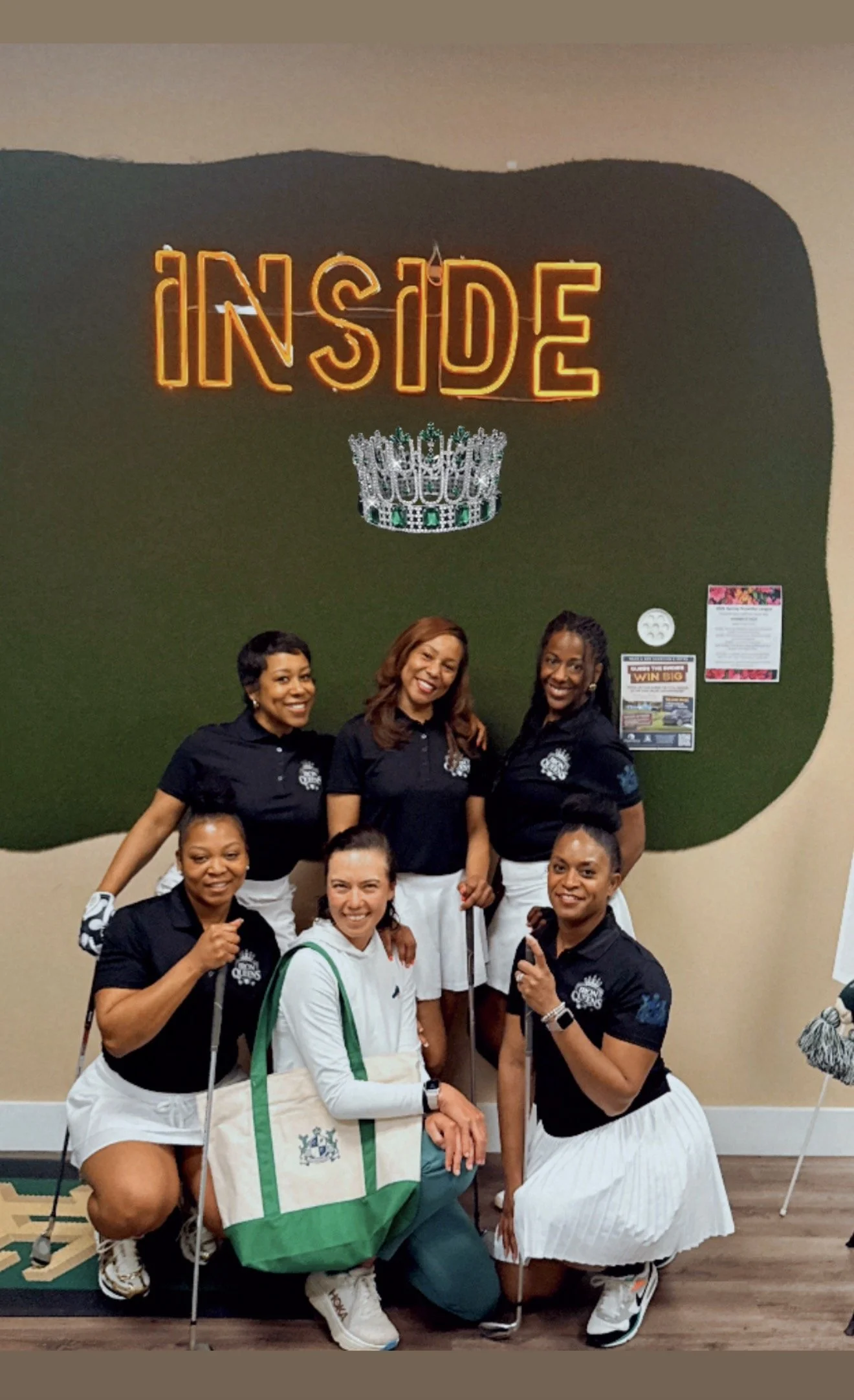Group of eight women in black and white golf attire, smiling and posing indoors in front of a green wall with neon sign reading 'INSIDE' and a decorative crown