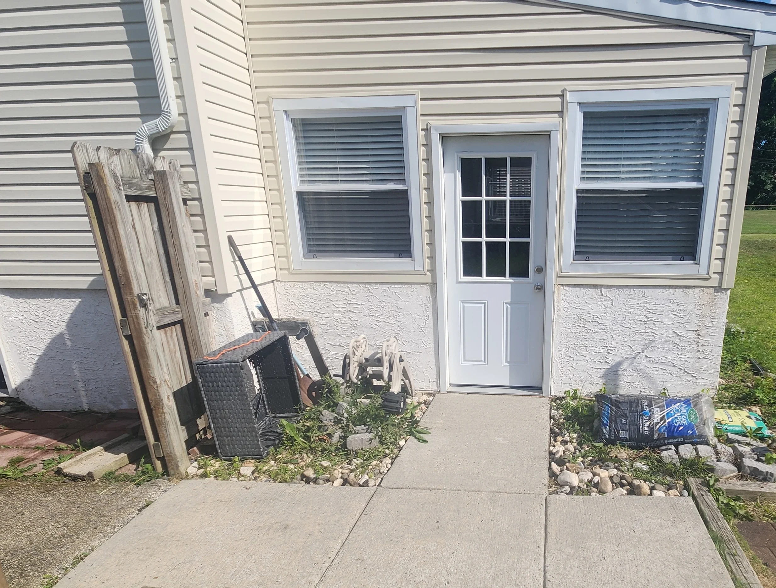 Backyard scene with a house door and two windows, construction materials, a wooden ladder, a black outdoor chair, bags of soil or mulch, and a small patch of grass and rocks. Looks cleaner after being pressurewashed.