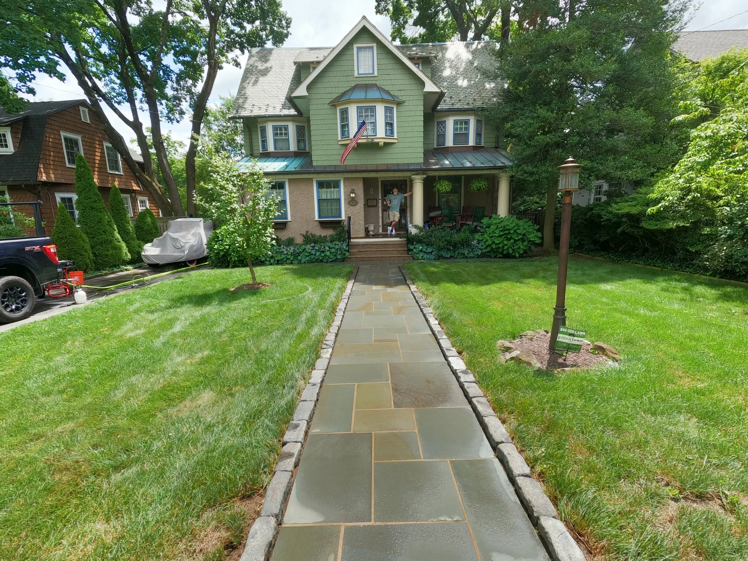 Front yard view of a two-story green house with a porch, trees surrounding, a man standing on the porch, and a sidewalk leading to the front steps. This is after cleaning.